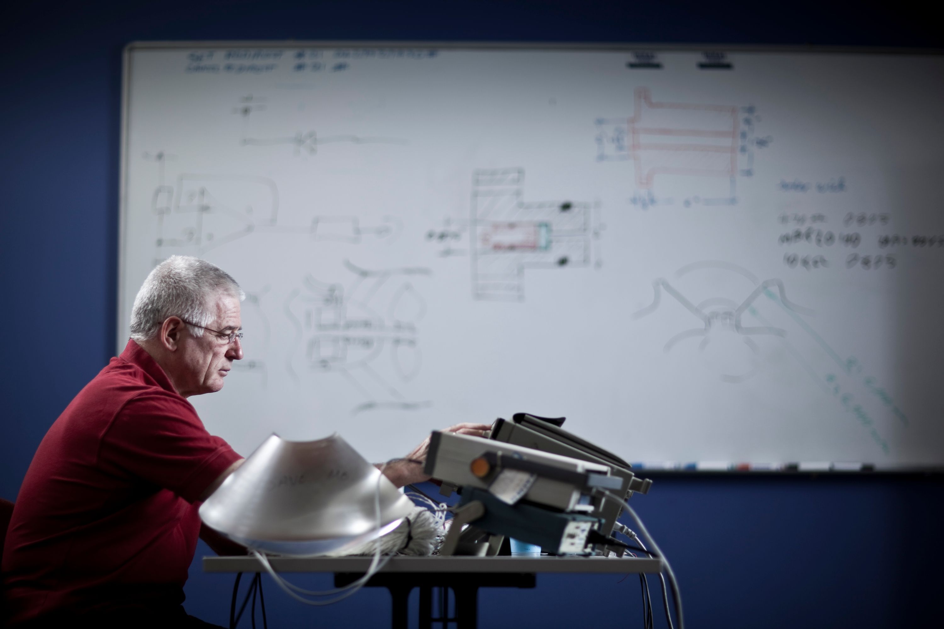 A man with white hair, glasses and a red shirt is seated at a table at the bottom left of the image. There is an out of focus whiteboard in the background.