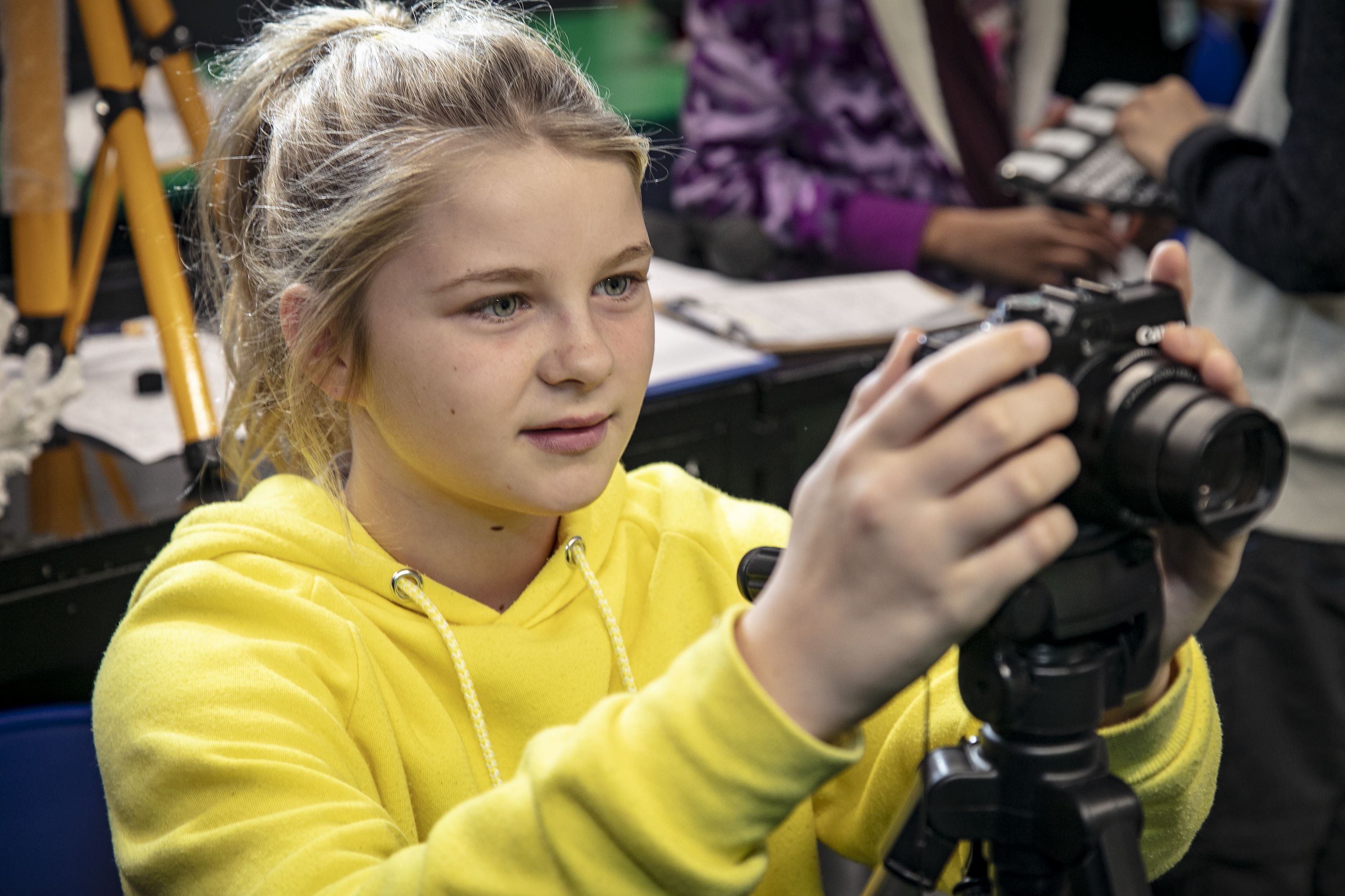 Photo of a girl with blonde hair and a yellow hoodie adjusting a camera on a tripod.