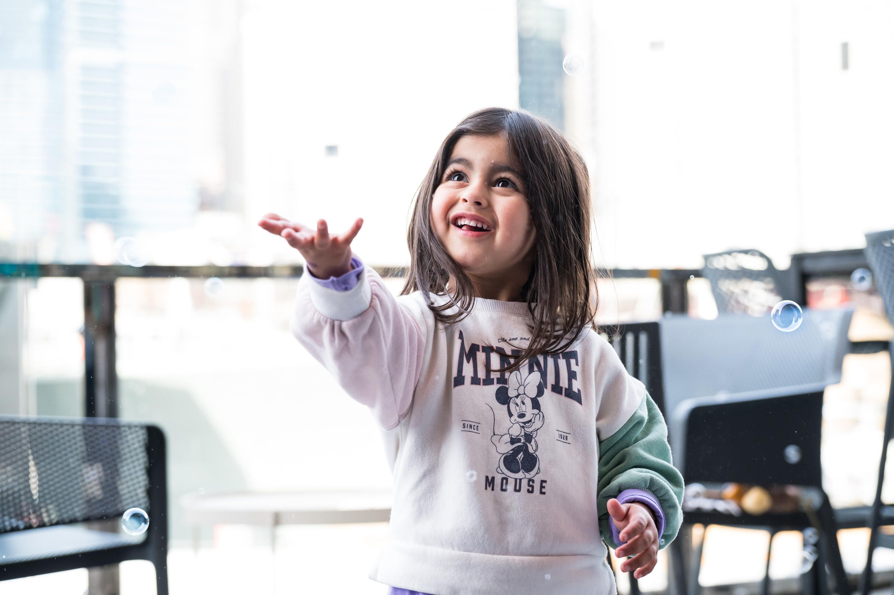 Photo of a young girl smiling and reaching her hand forward to catch a bubble out of the air. 