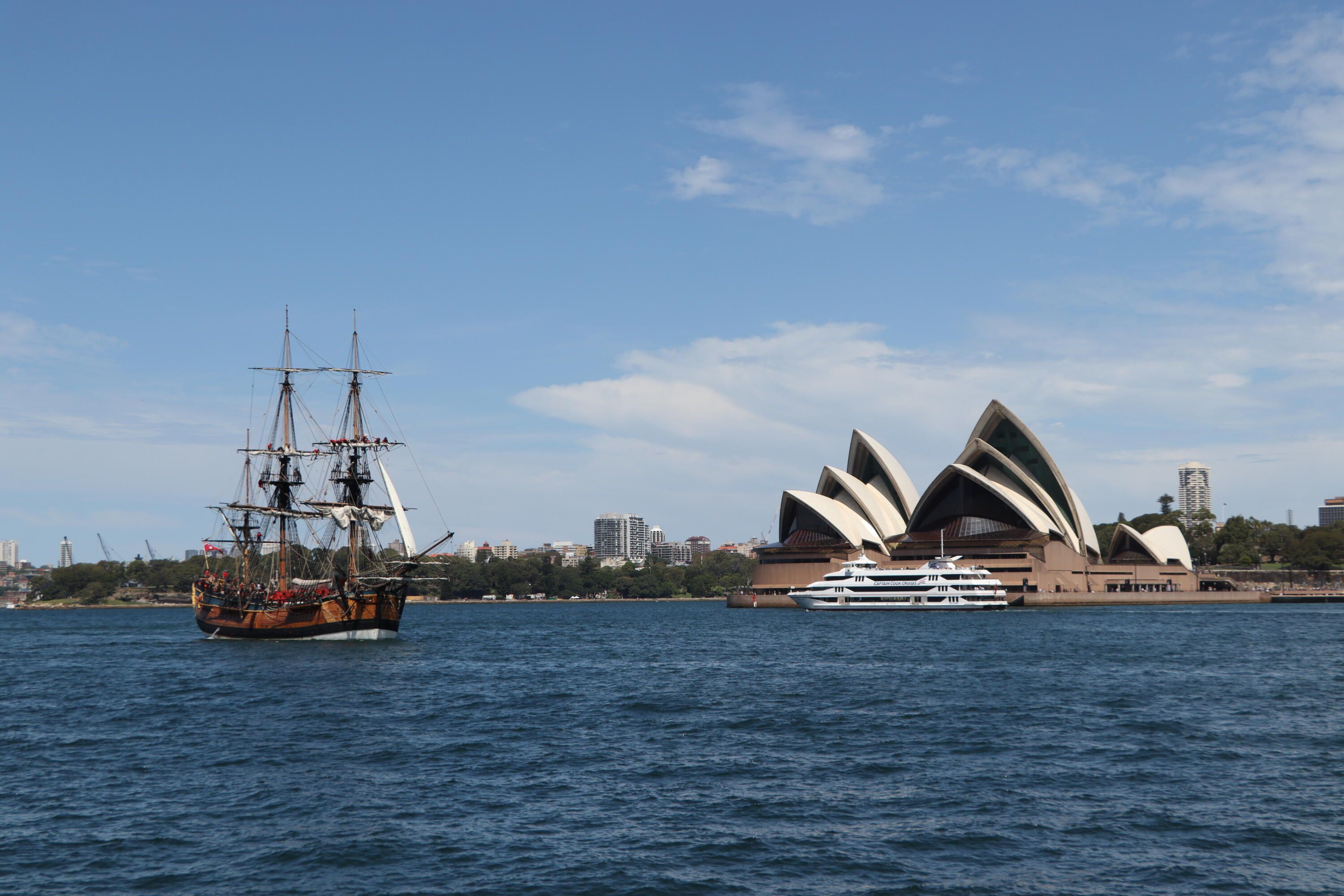 Photo of a tall ship, Endeavour, its sails furled, sailing in Sydney Harbour, with the Sydney opera house and a white ferry to the right of it. 