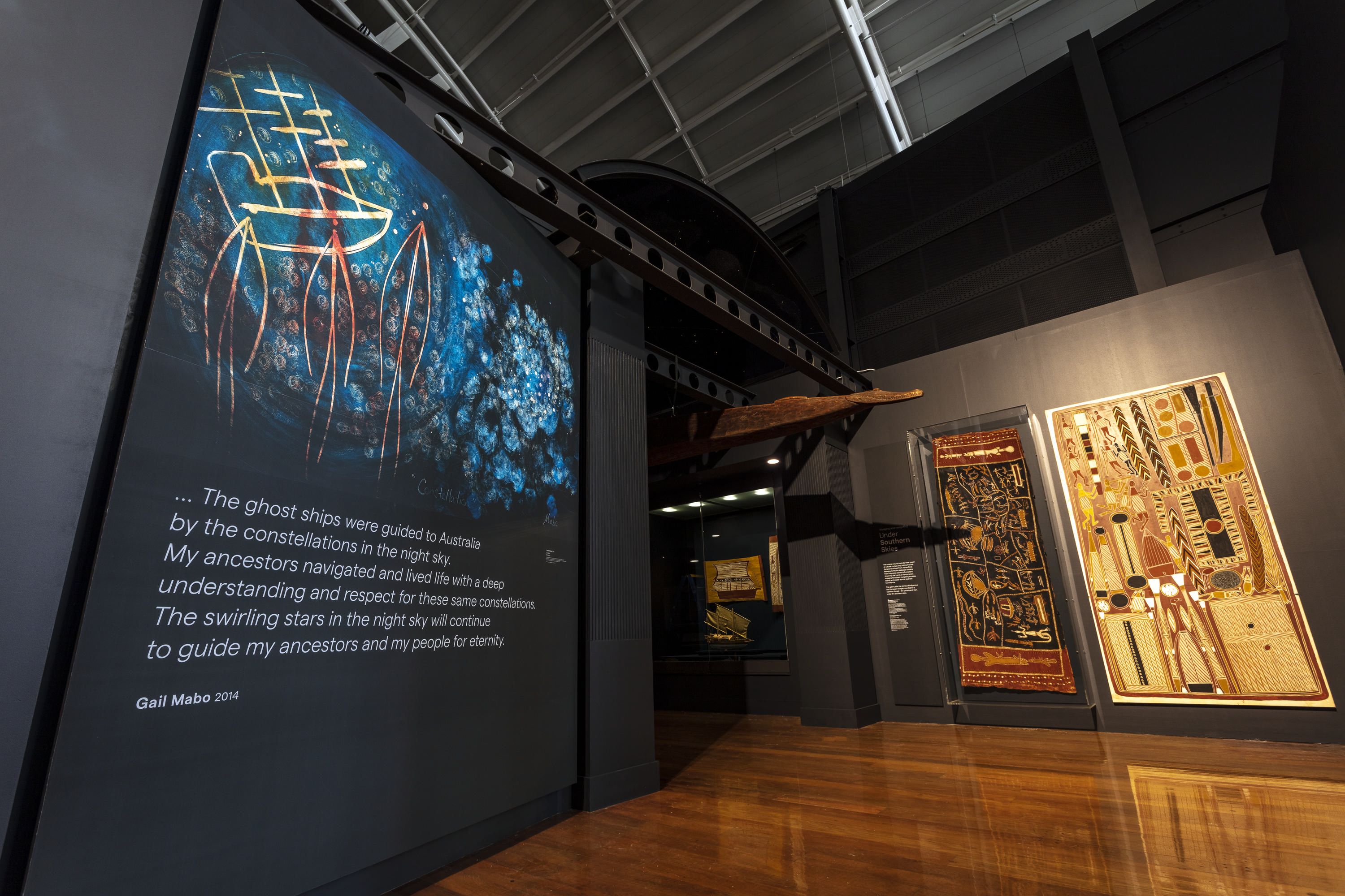 Photo of the entrance to a museum exhibition. There is a entry wall featuring a contemporary first nations artwork and text, and bark paintings on the opposite wall. A crocodile carving is hanging from the celling. 