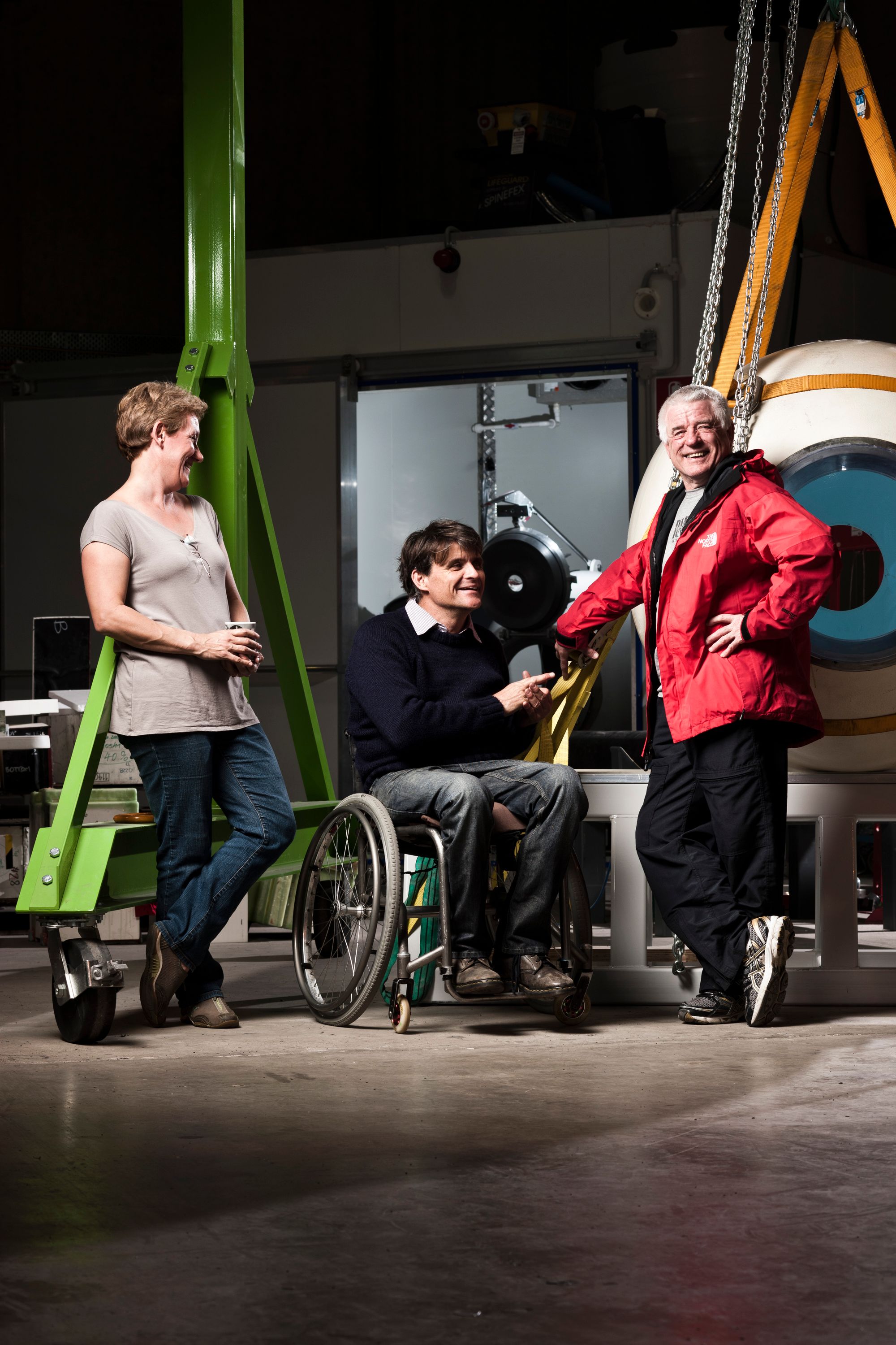 A group of three people posed for a photo; A woman with short hair a tan top and jeans, a man with dark hair and a black jumper sitting in a wheelchair, and a man with white hair and a red coat.