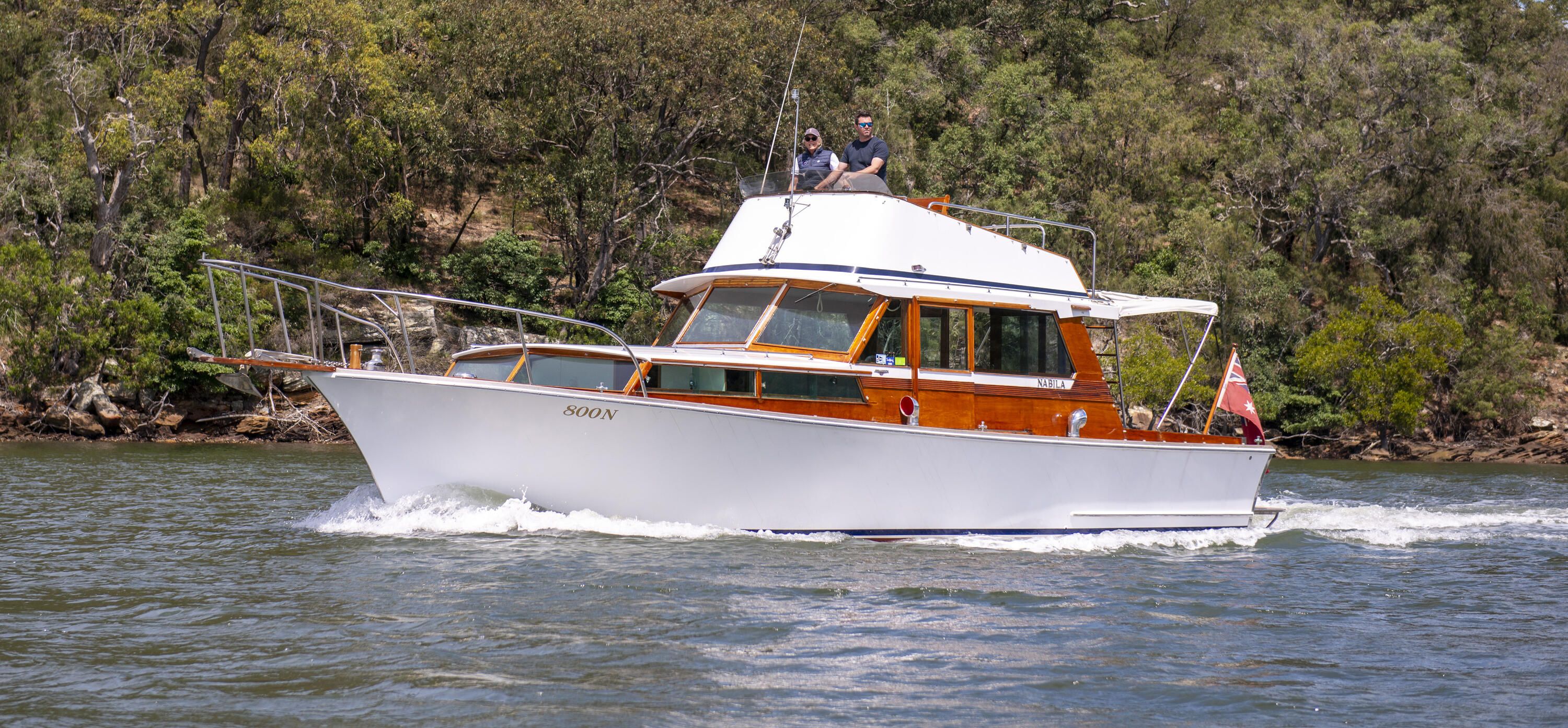 hoto of a boat with a white hull and wooden features, driving on the water with green foliage behind it.