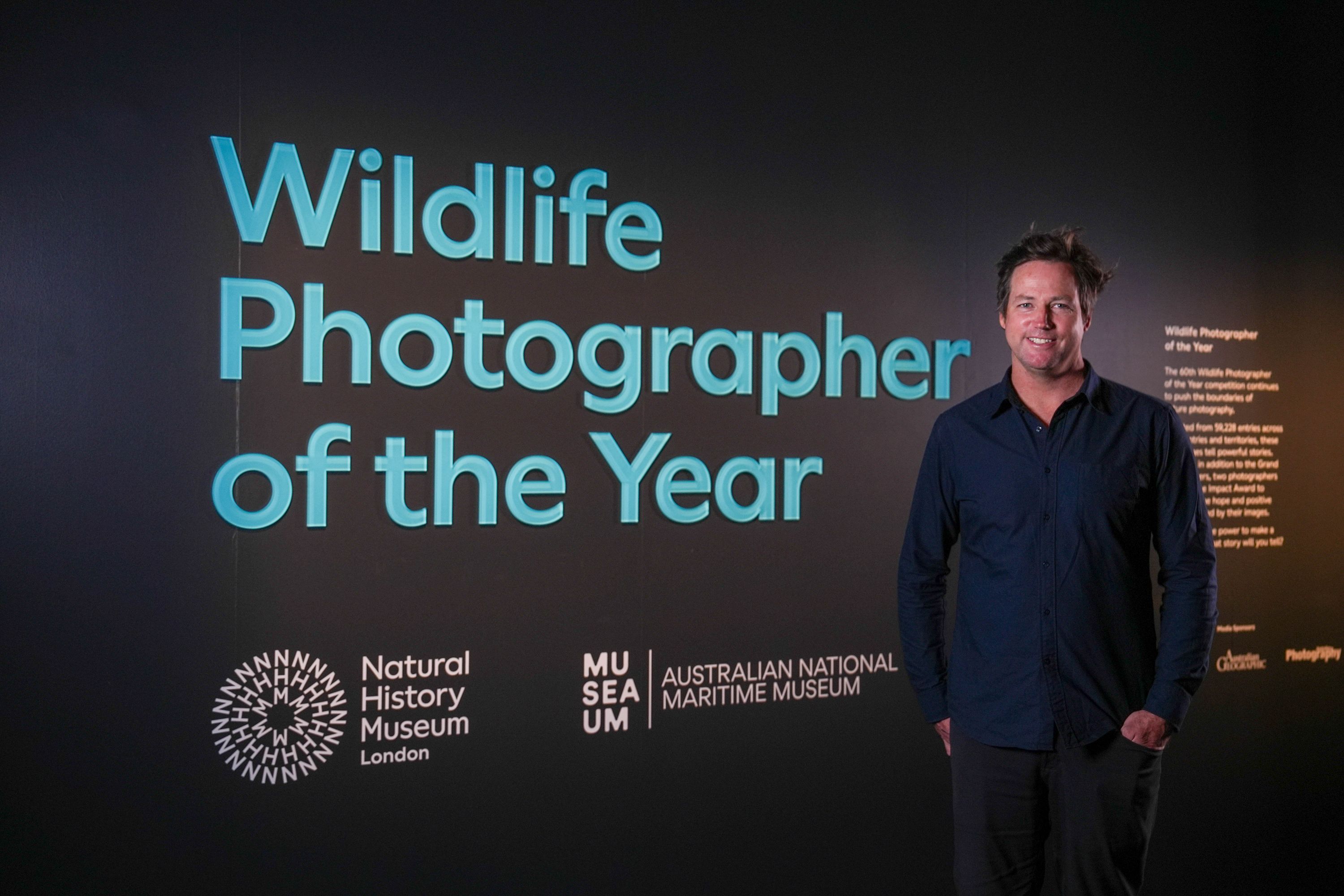 Photograph of an entry wall to an exhibition called Wildlife Photographer of the Year with a man standing in front of it.