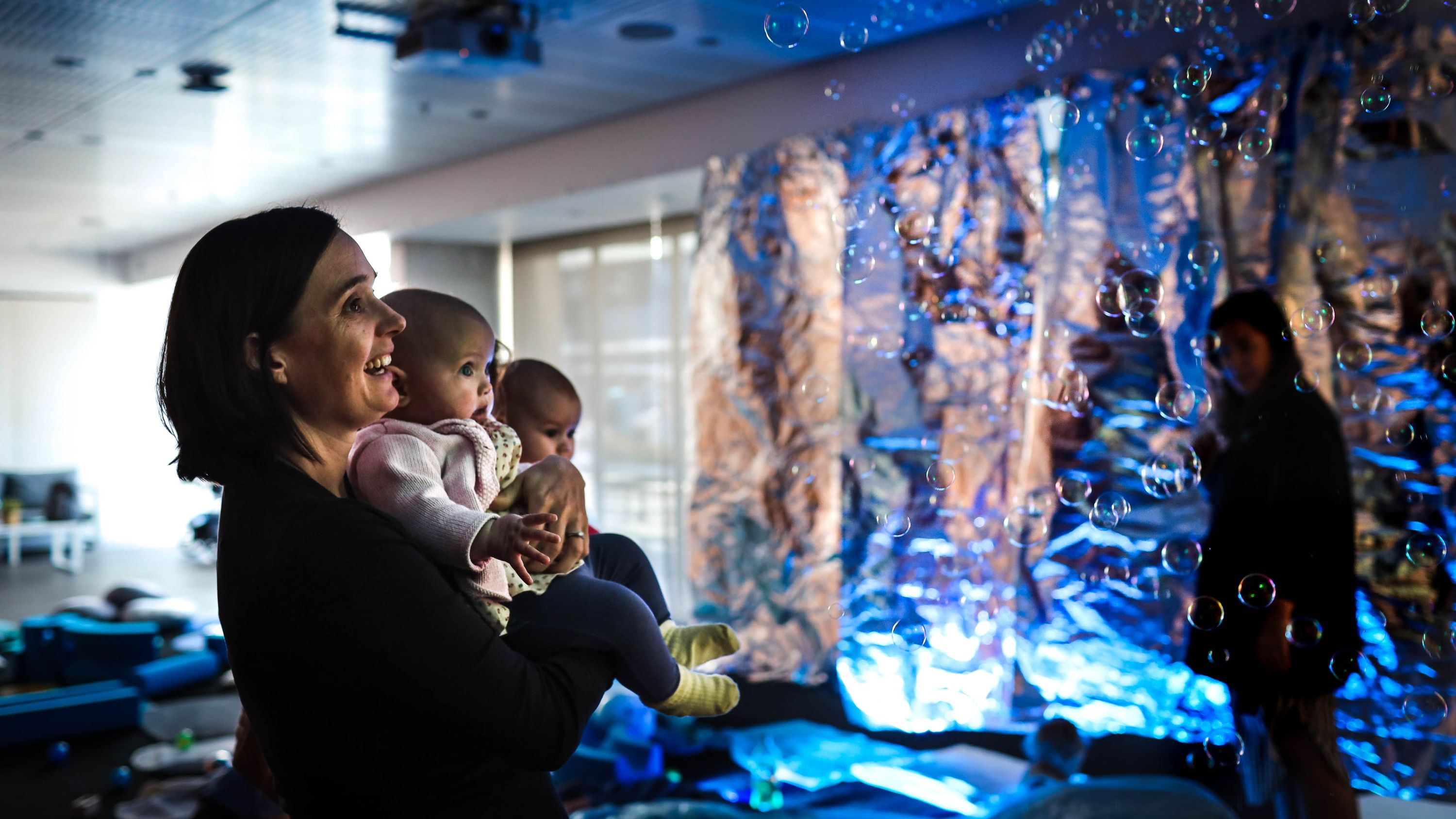  Mums and babies in the play space. A mum and bub watch floating bubbles drift by them.