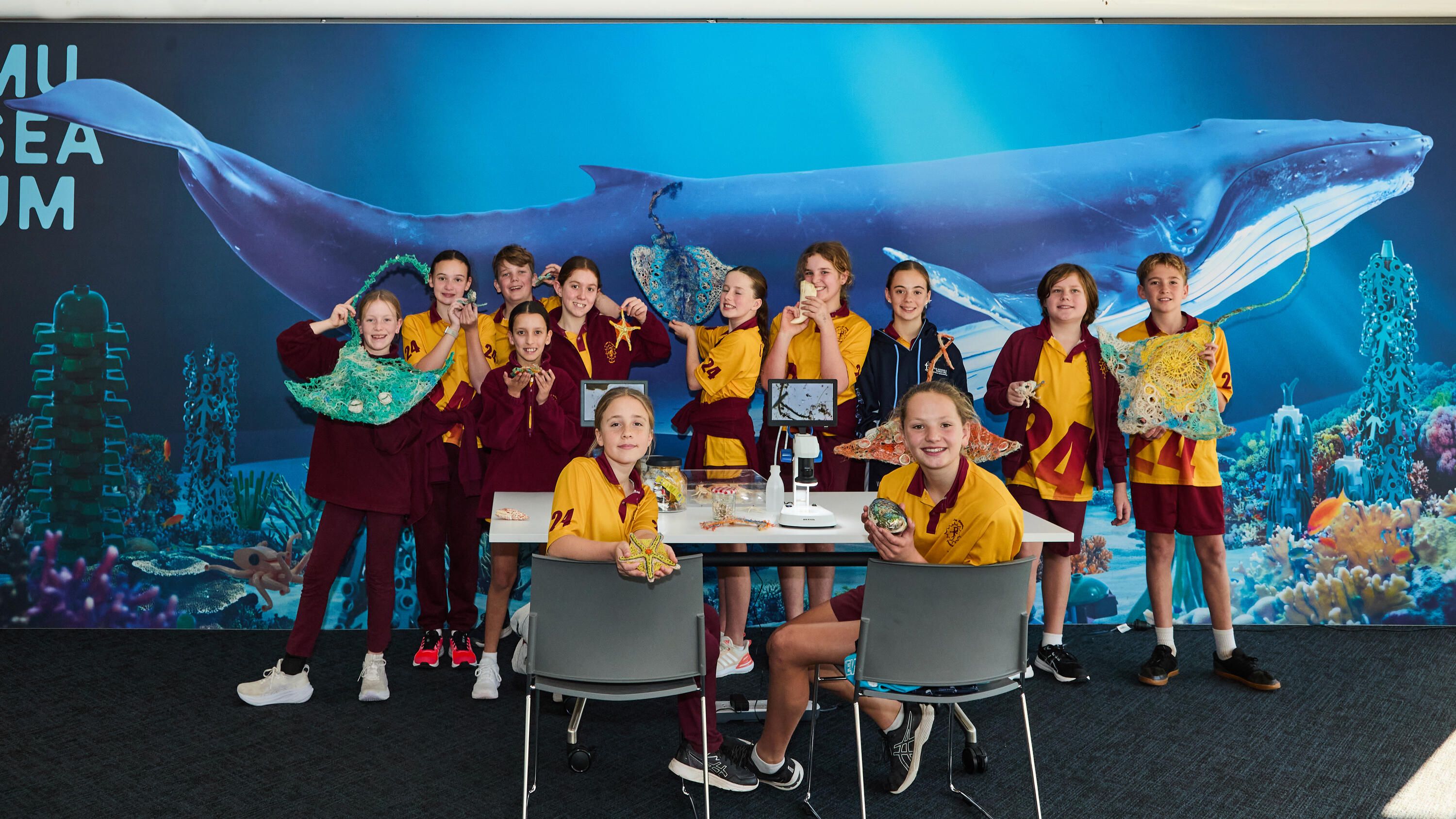 Photo of a group of primary school students in uniform standing around a table with an illustration of a whale in the background.  