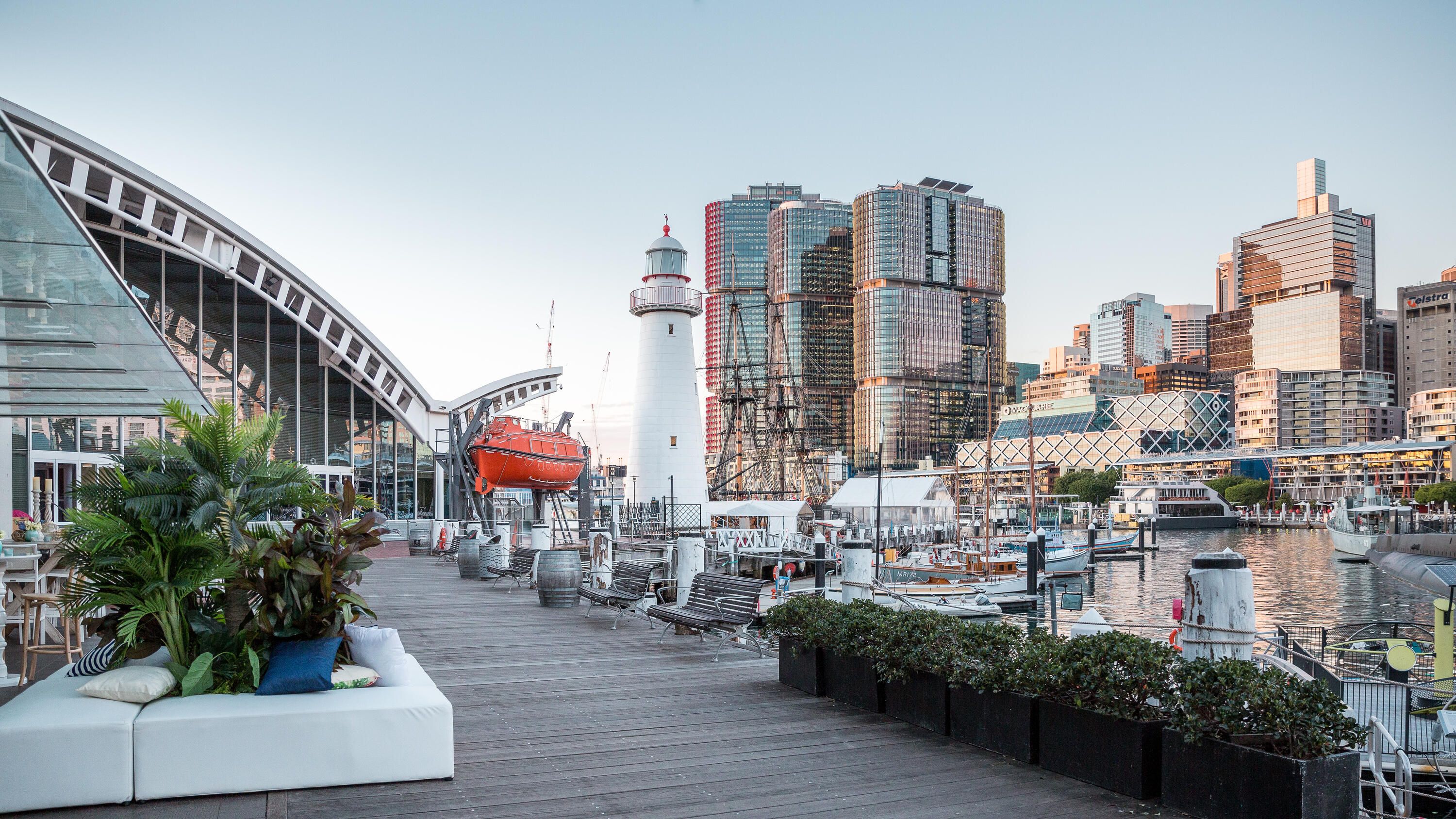 photo of outside the museum, with the boardwalk in the foreground, and a lighthouse and the Sydney skyline in the background on a sunny day. 