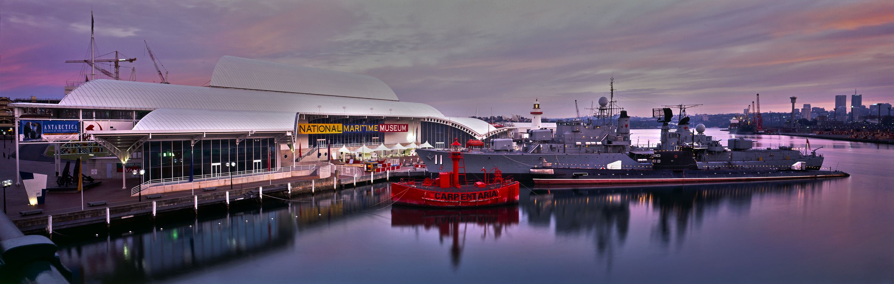 Photograph at sunset with a pink sky, showing the white museum building, with various vessels in the foreground. 