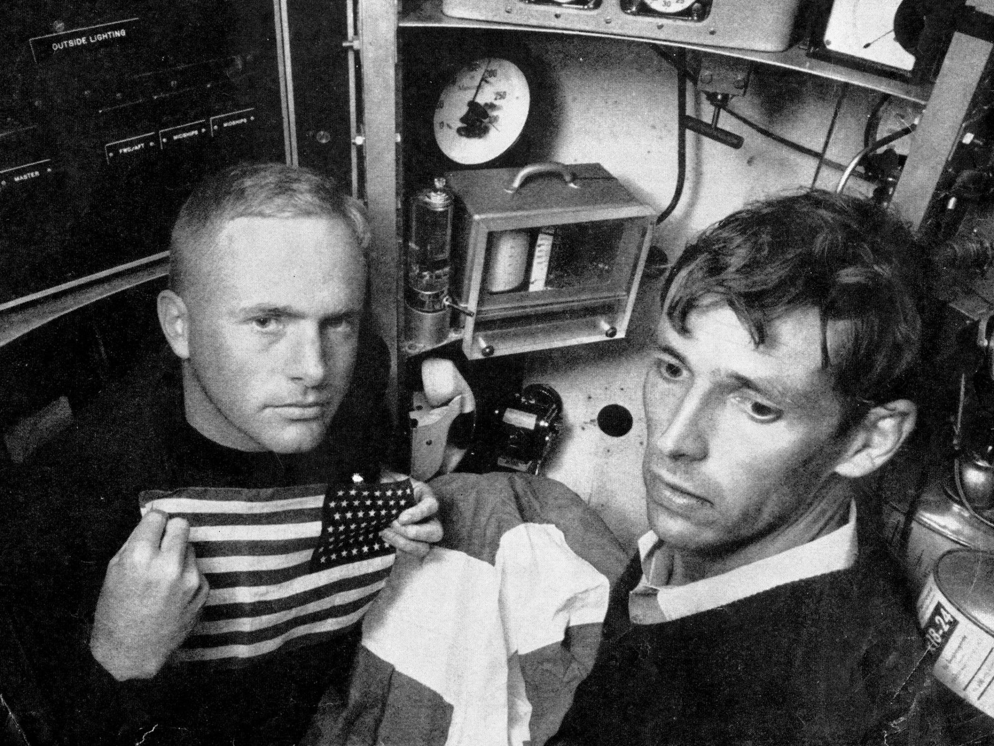 Black and white photo of two men inside a small space. One holds an American flag.