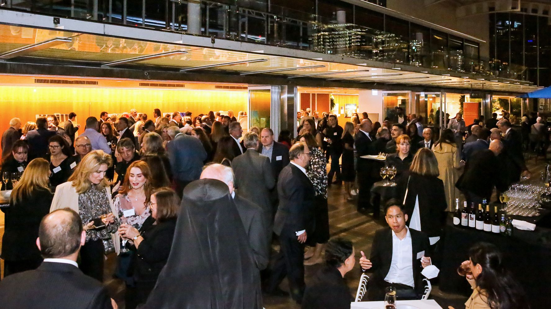 Photo of a crowd of well dressed people at an event, talking and drinking on the museum boardwalk, with the buildingin the background