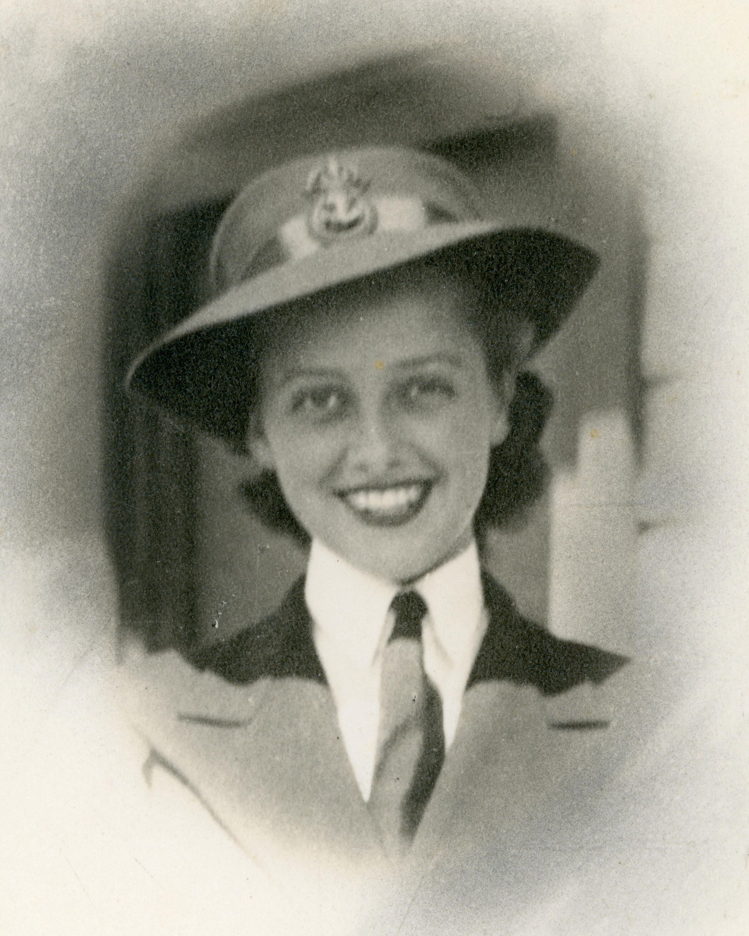 An old, black and white photo showing the face of a young woman, with short dark hair,  wearing a hat that features a badge in the centre, a white shirt, dark tie and dark jacket. 