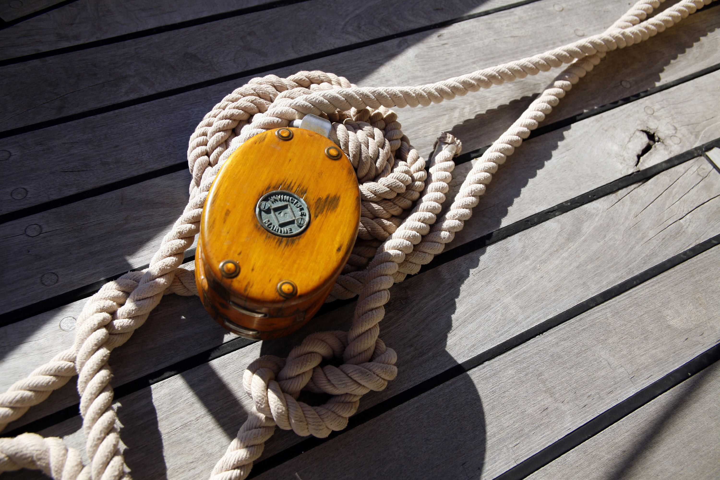 Close up photo of ropes, knots and a pulley in the sun on timber decking aboard a wooden boat. 