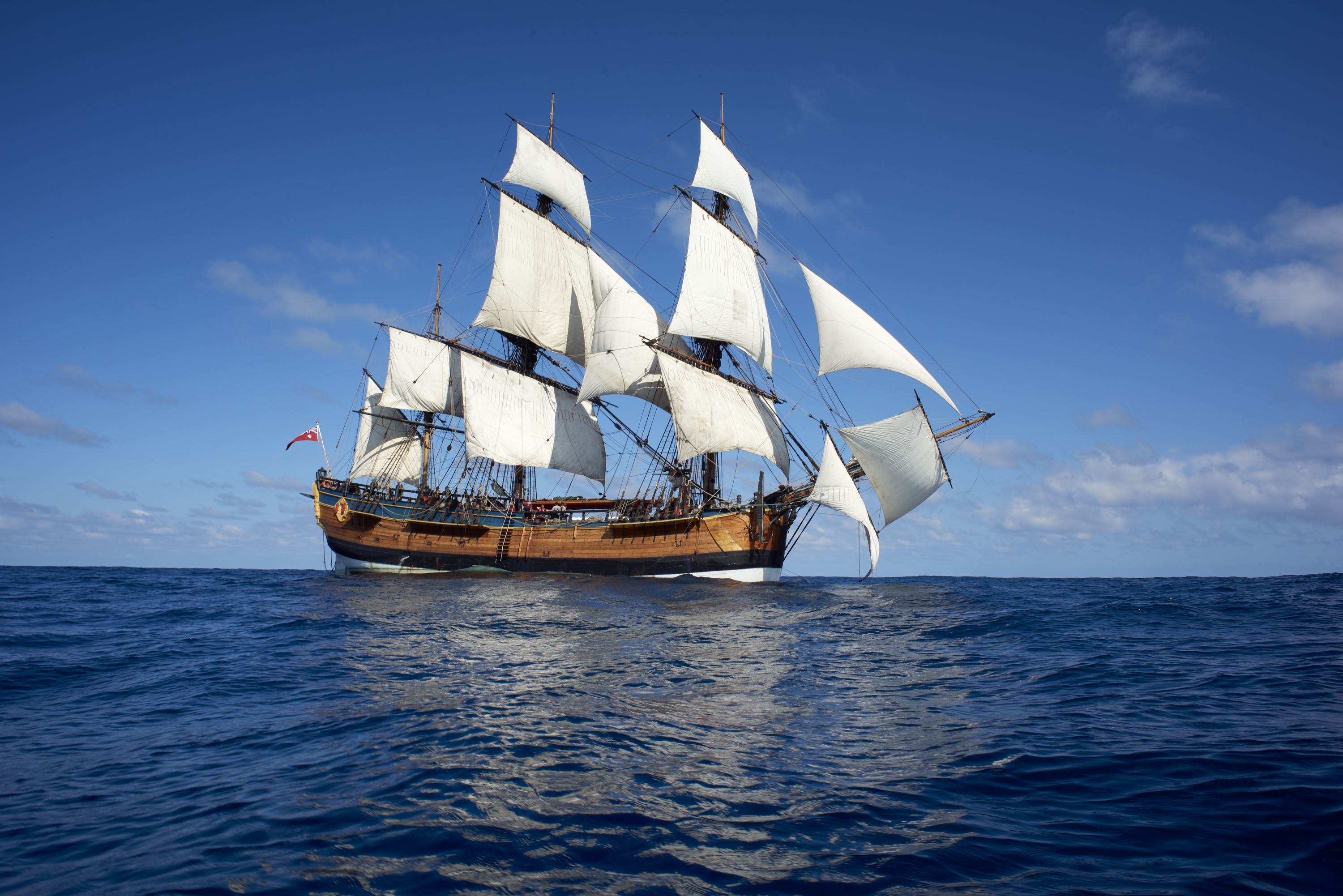 Photo of a tall ship with white sails on a bllue ocean with a blue sky. 