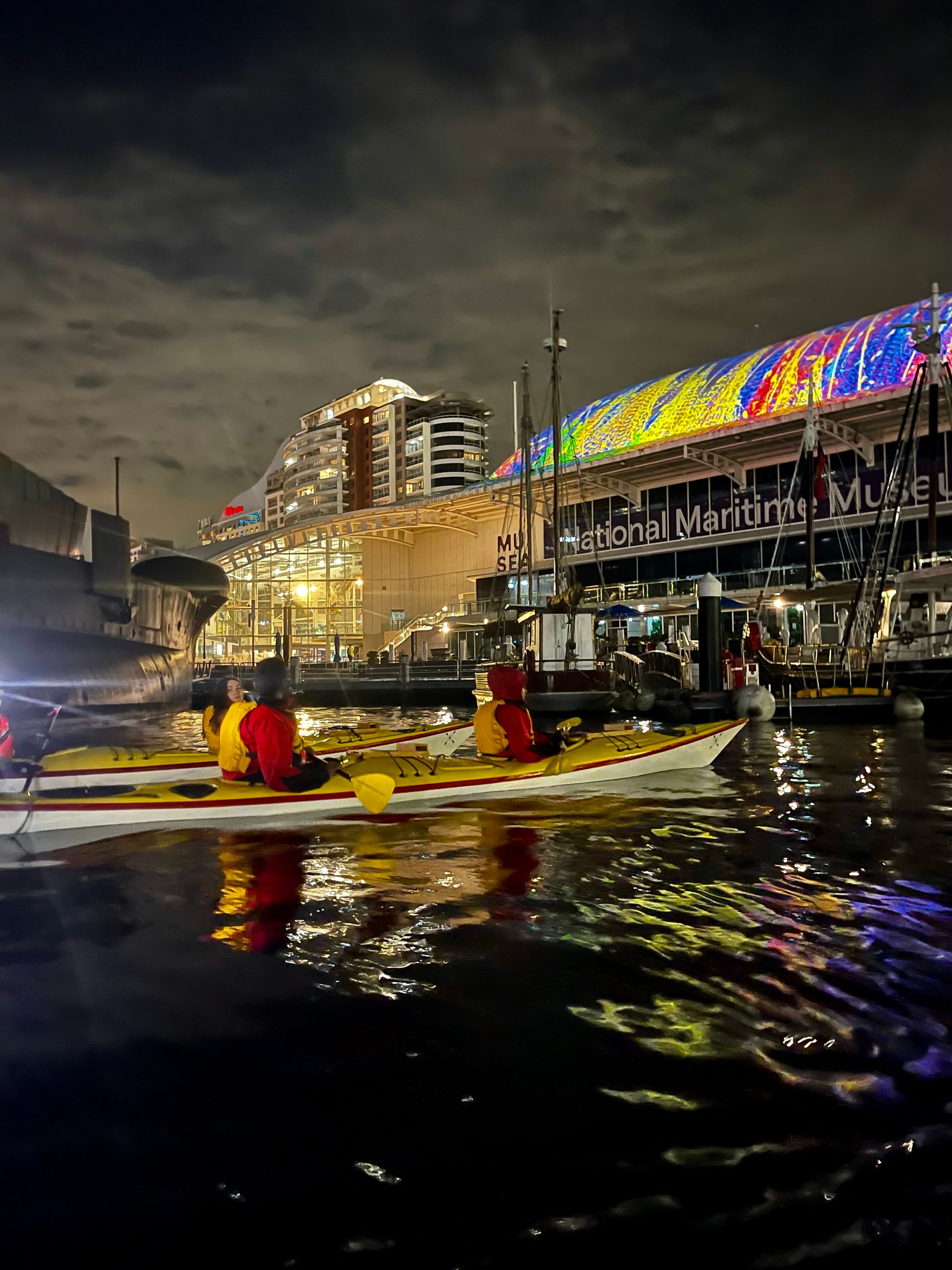 Photo taken at night showing 2 people in a double sea kayak infront of the brightly lit museum building with a colourful projection on the sloping roof. 