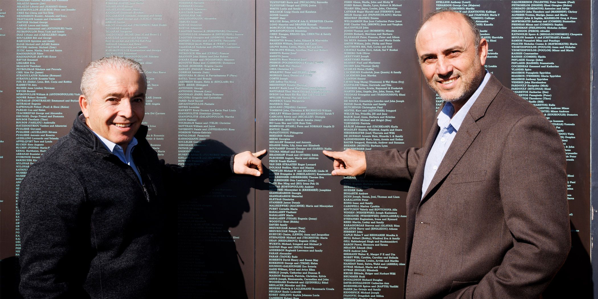 Photo showing 2 smiling men pointing at a list of names engraved in a metal wall. 