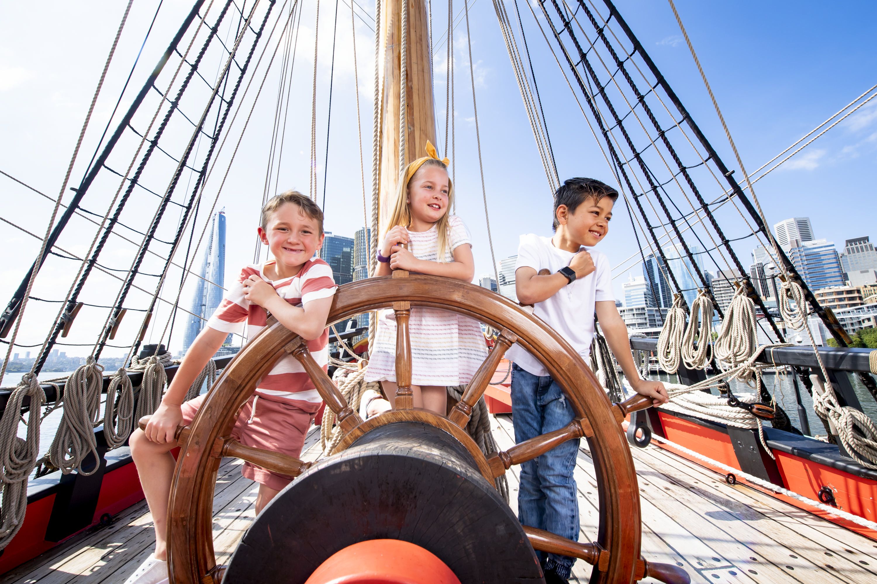 Photo showing two boys and a girl on board a tall ship and pose behind the ship's wheel.