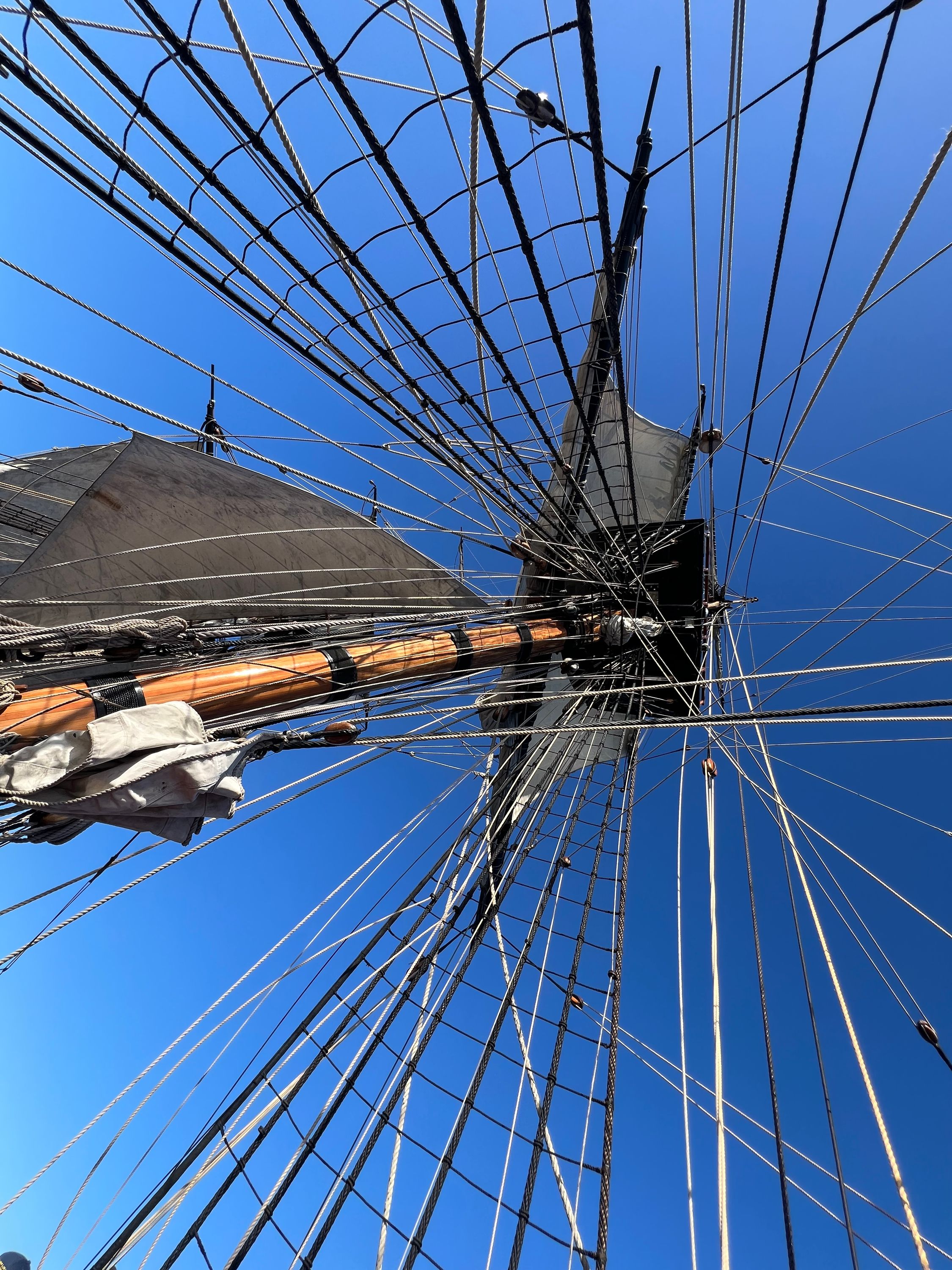 A view looking up at a tall ship's mast, with many ropes leading up. The blue sky is behind it. 