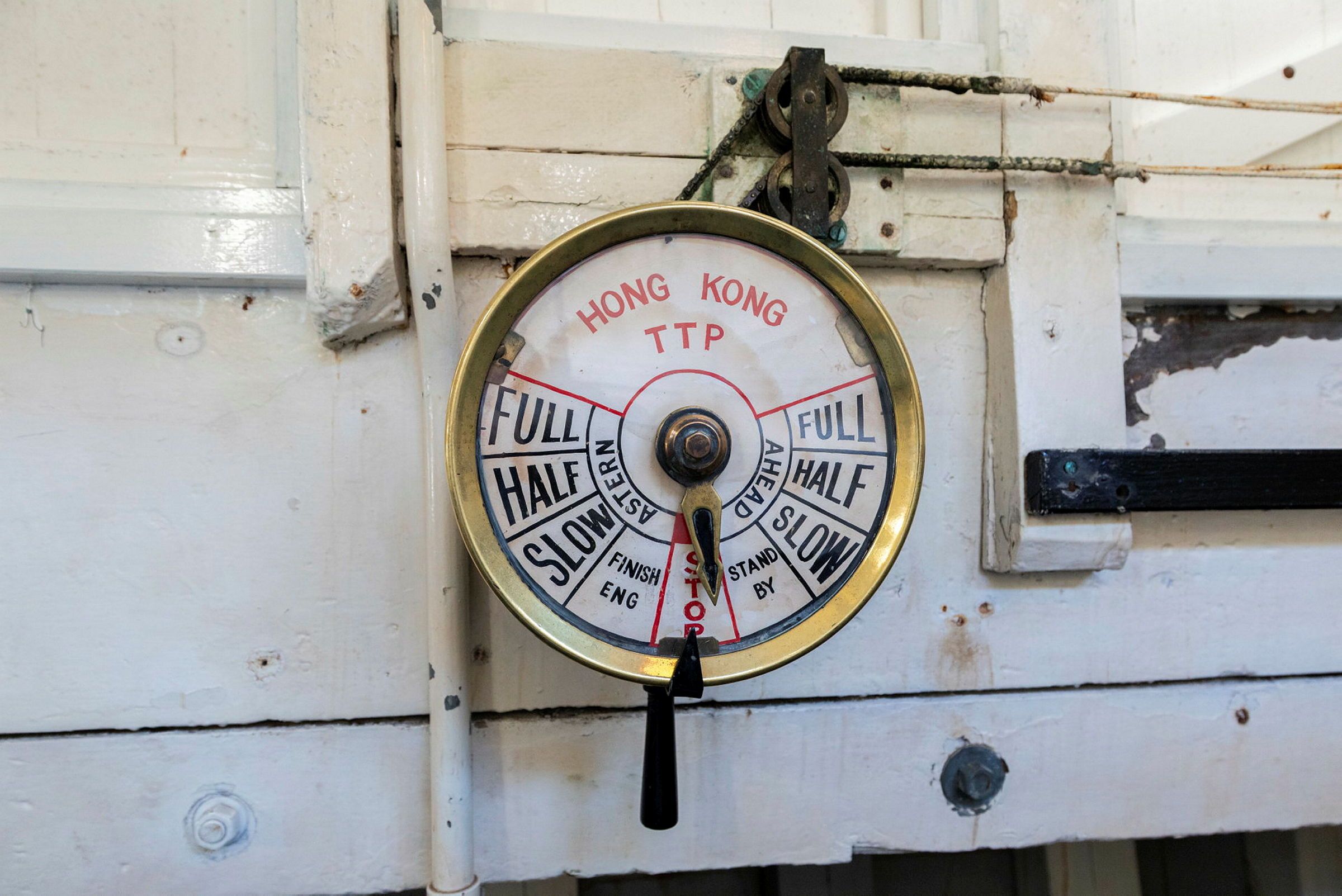 Photograph showing a ship's telegraph. A round face with writing on it such as full, half and slow, with a hand pointing towards different speeds.
