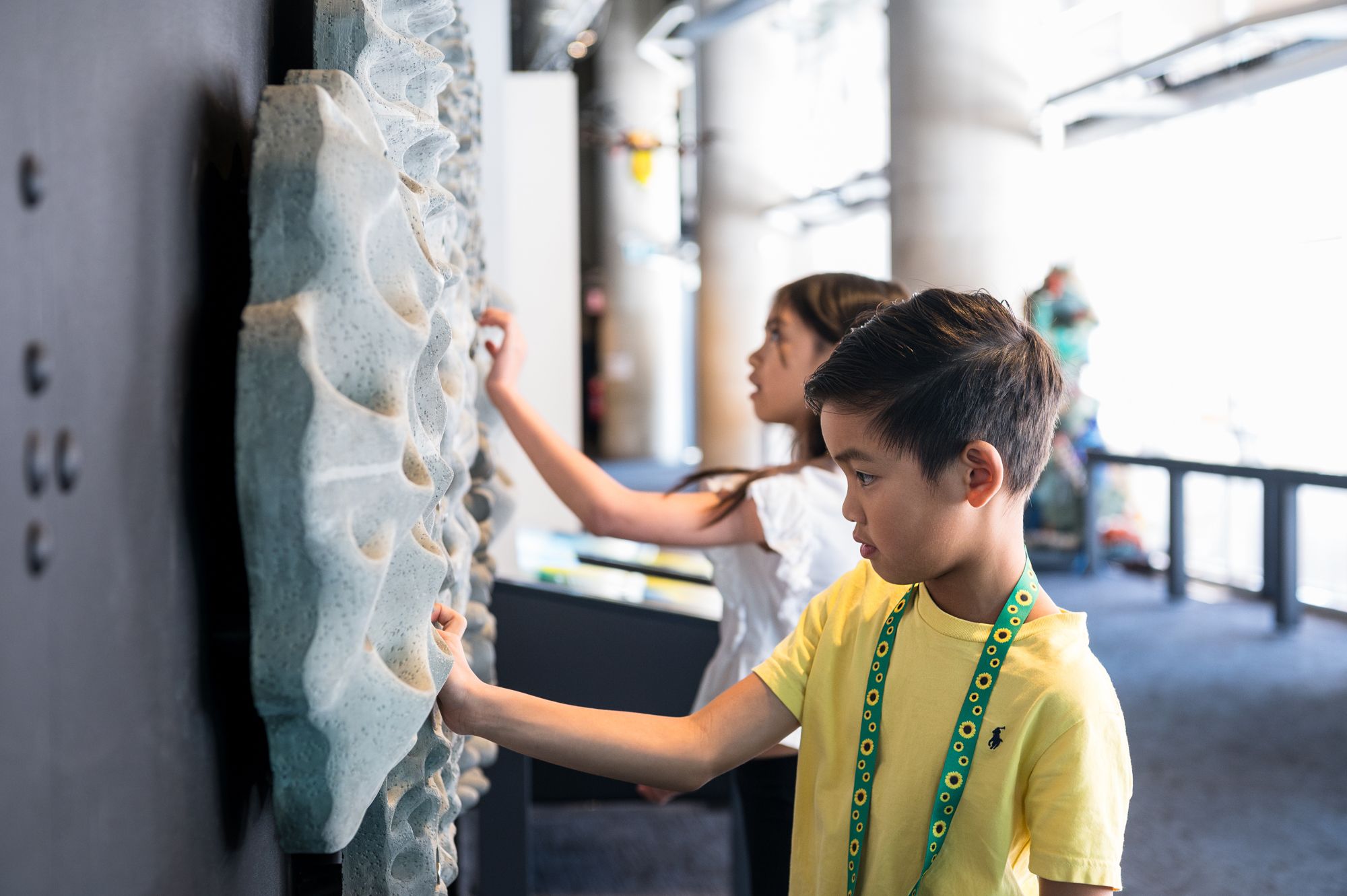 Young boy with short, dark hair, wearing a yellow shirt and sunflower lanyard touching a sculpture on the gallery wall, with a young girl out of focus behind.