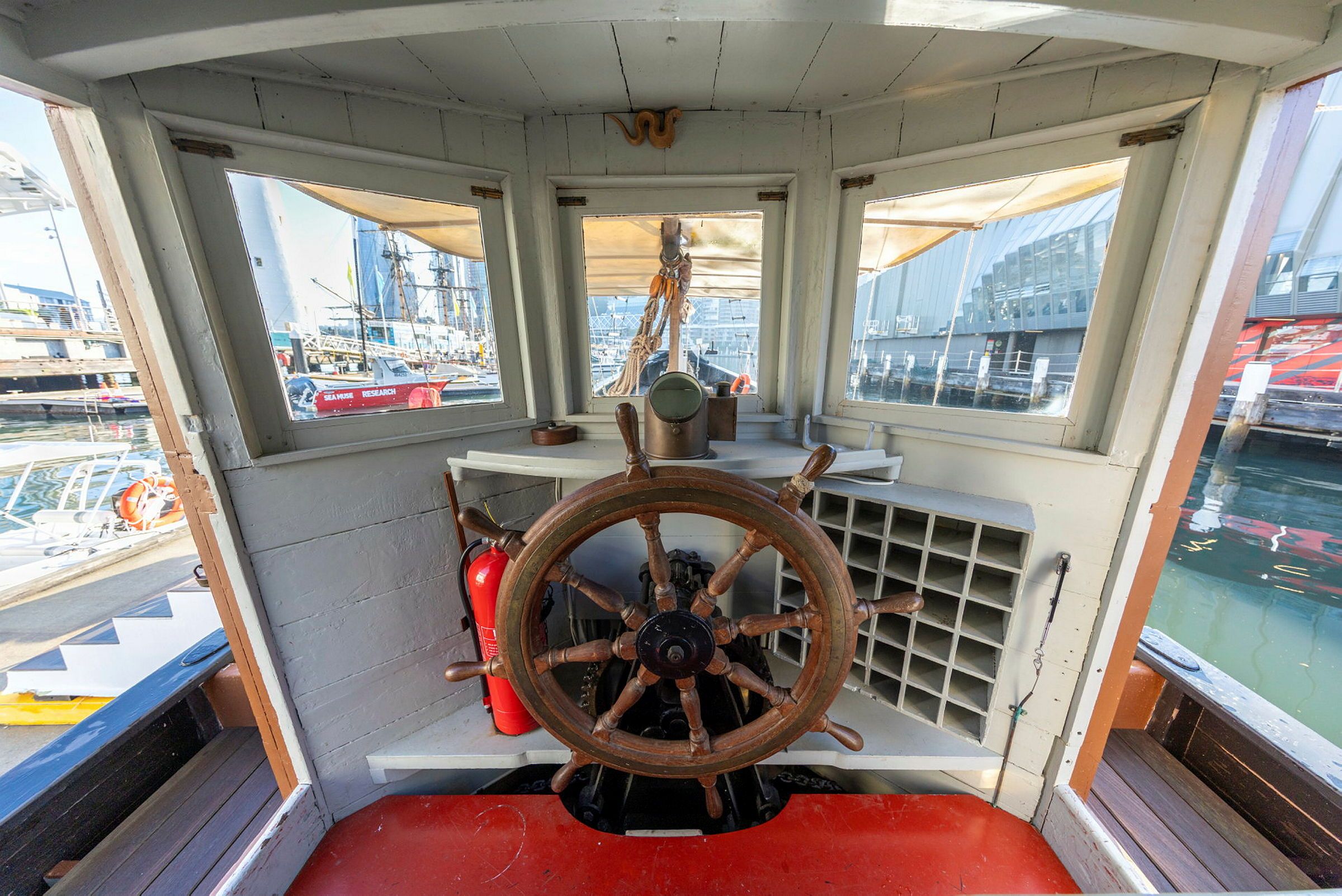 Photo taken inside a small cabin on a boat, looking at a large ship's wheel. 