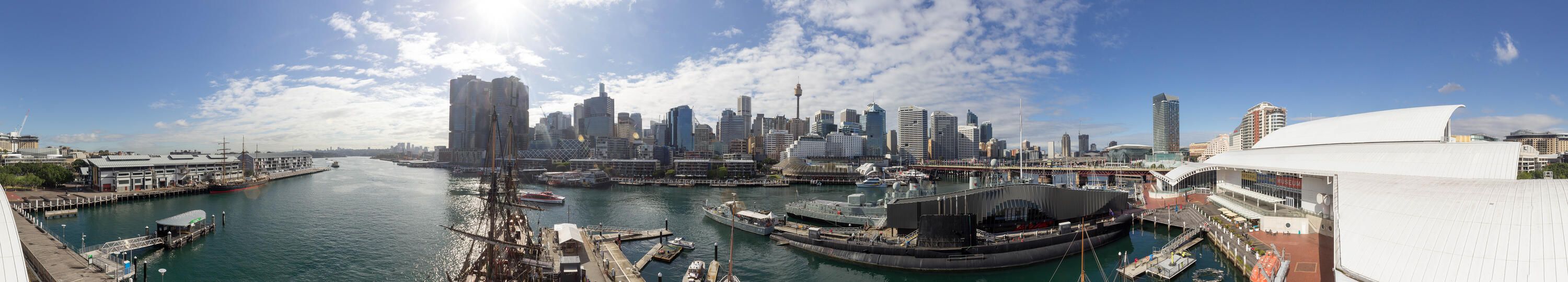 Panorama showing Sydney city and Darling Harbour on a bright sunny day with blue sky and clouds. There are wharves and various boat at the bottom, the blue green harbour and skyscrapers behind.