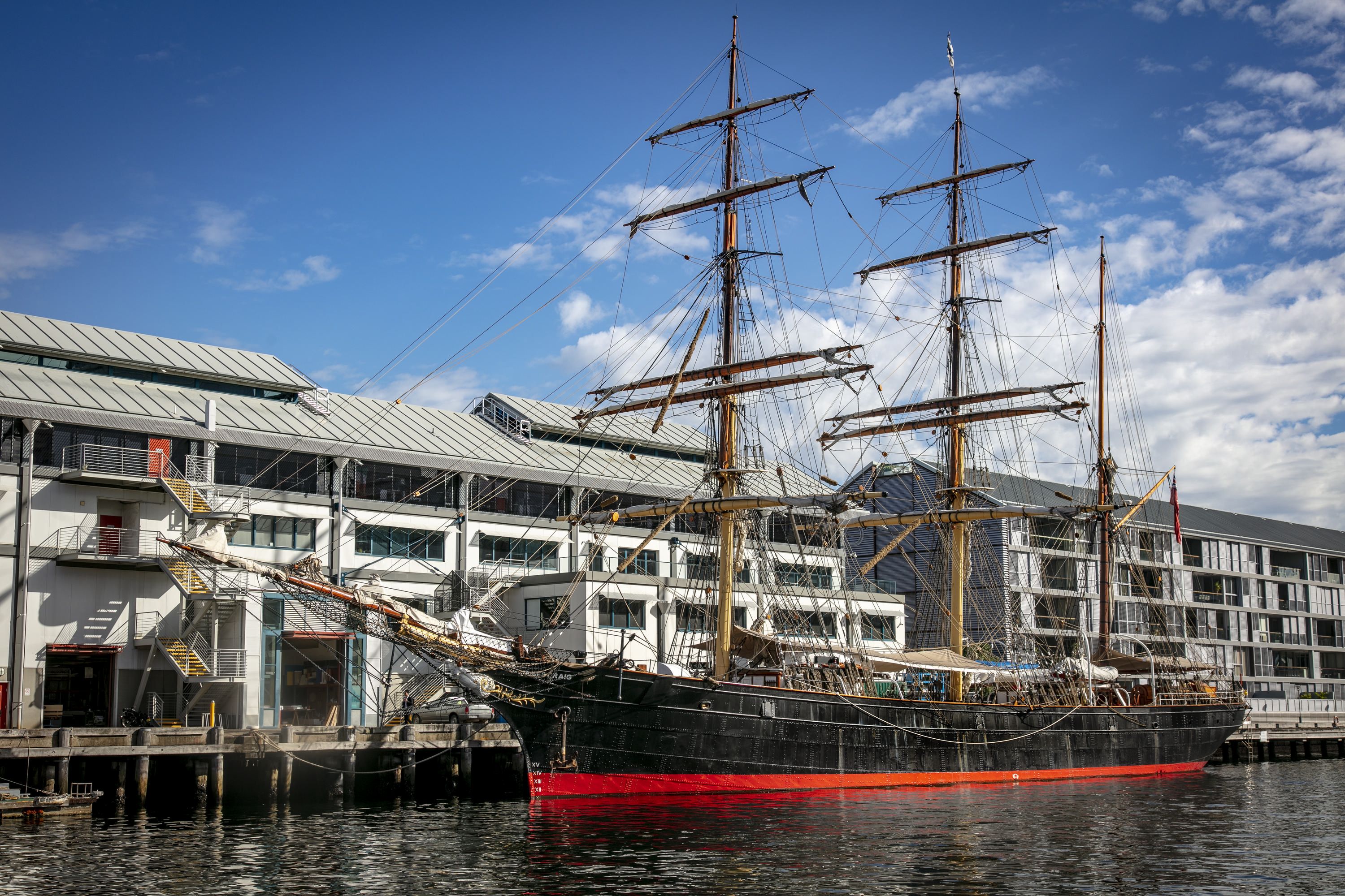 Photograph showing a tall ship with a black and red hull and 3 masts, anchored alongside a wharf with buildings behind it. 