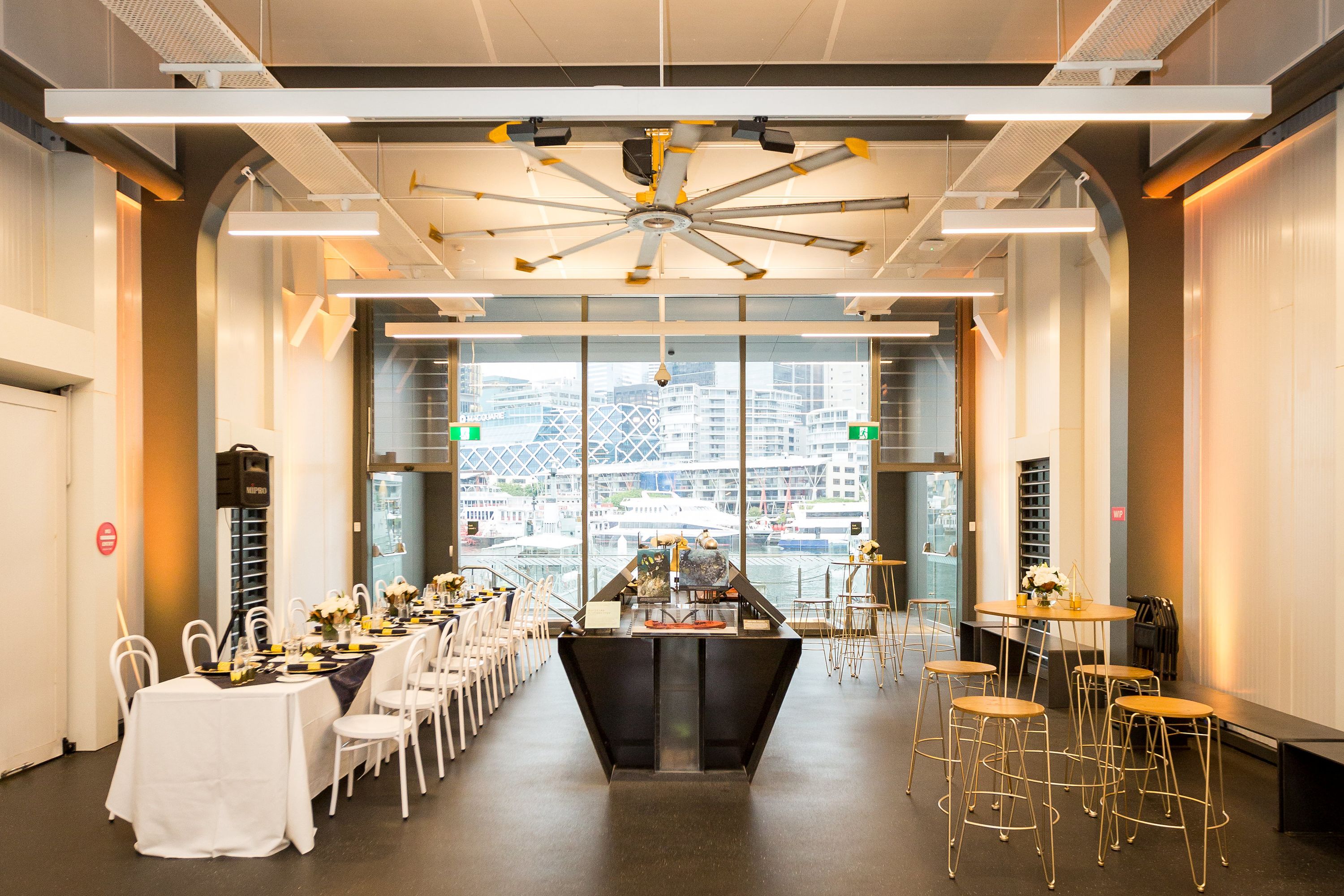 photo of a museum space with a long table set and decorated on the left side, museum interactive displays in the middle and a cocktail table and stools on the right, with a large window to the rear.