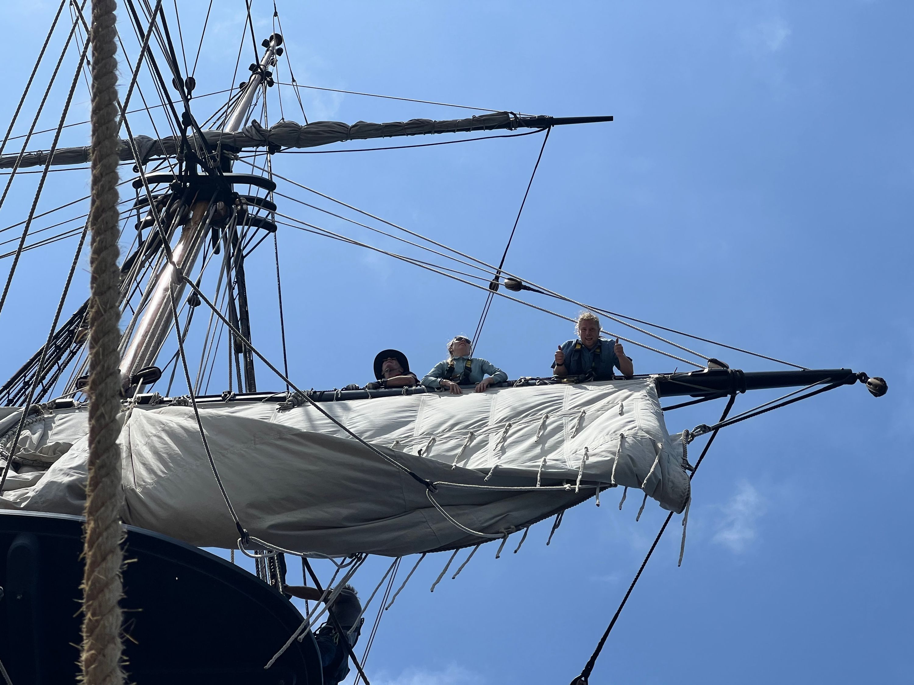 A photo taken up at a ship's mast and sail as 3 people are standing along the yard. 