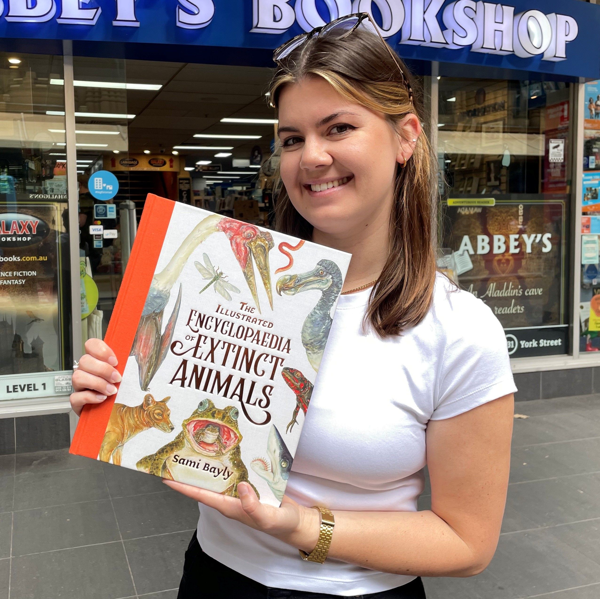 Photo showing a young woman standing outside a bookshop, holding a book called The Illustrated Encyclopaedia of Extinct Animals
