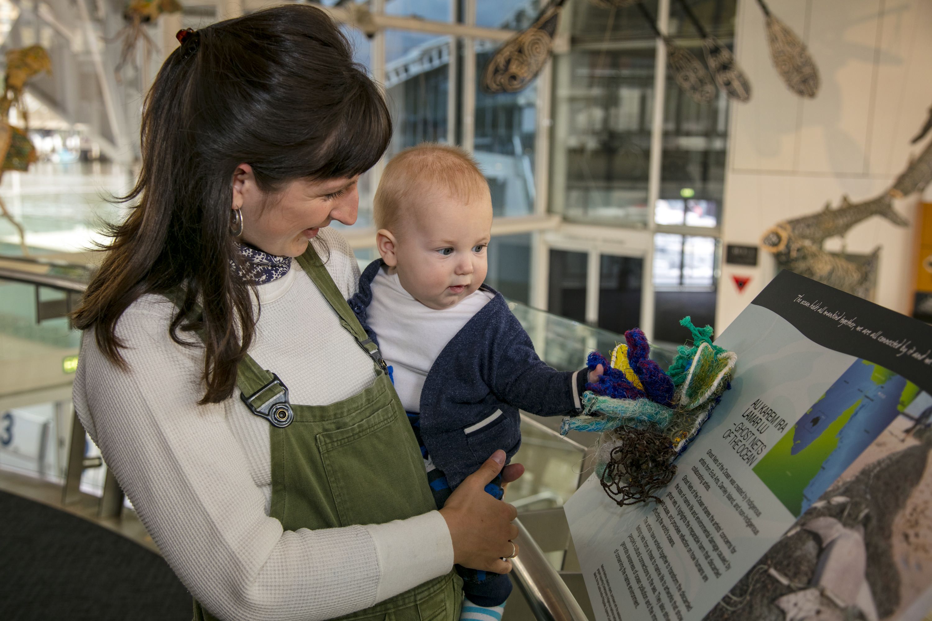 A new young mum, posed holding her baby in the museum with the Ghost Nets foyer display in the background.