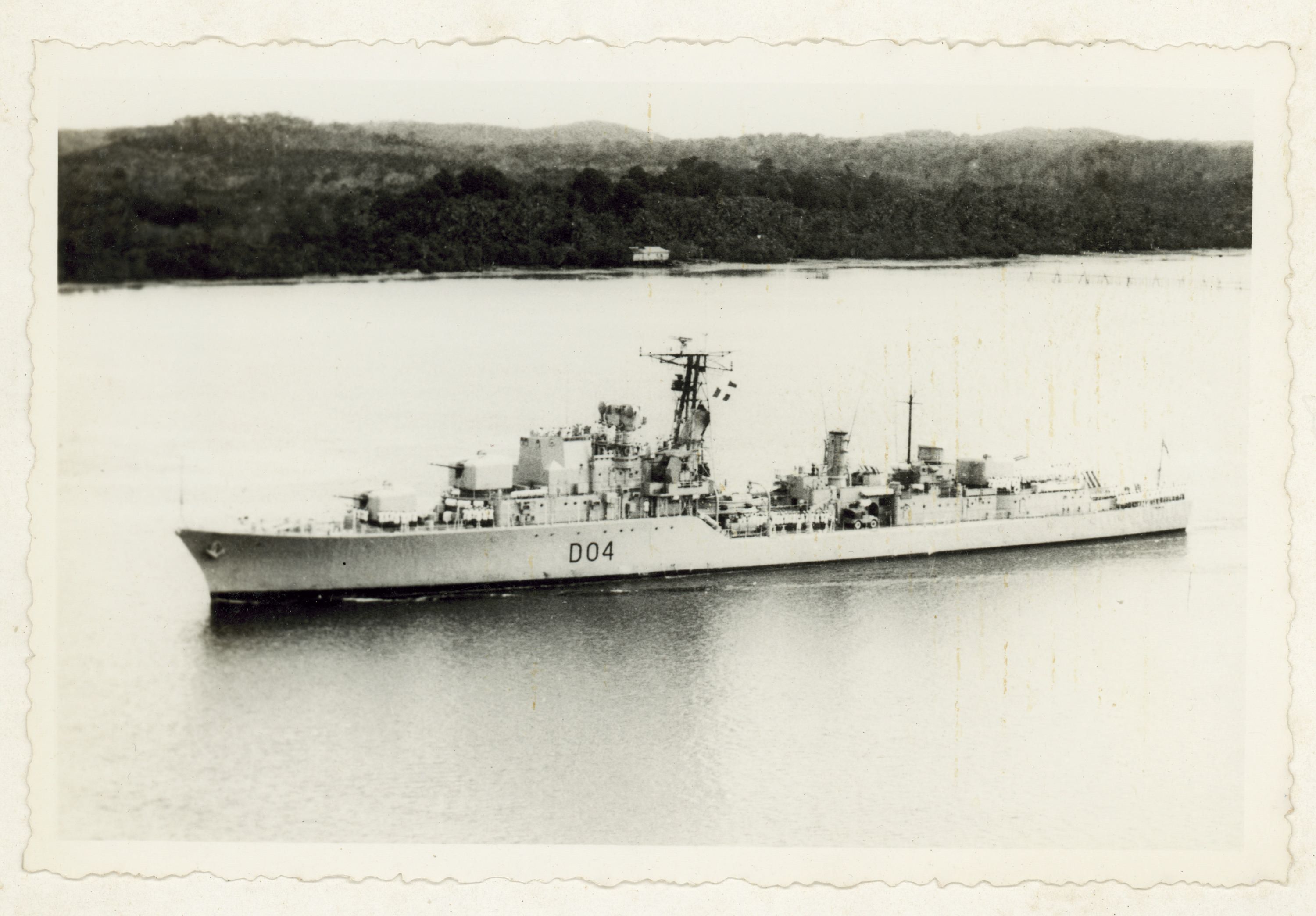 Black and white photograph showing a navy destroyer with land behind it. 