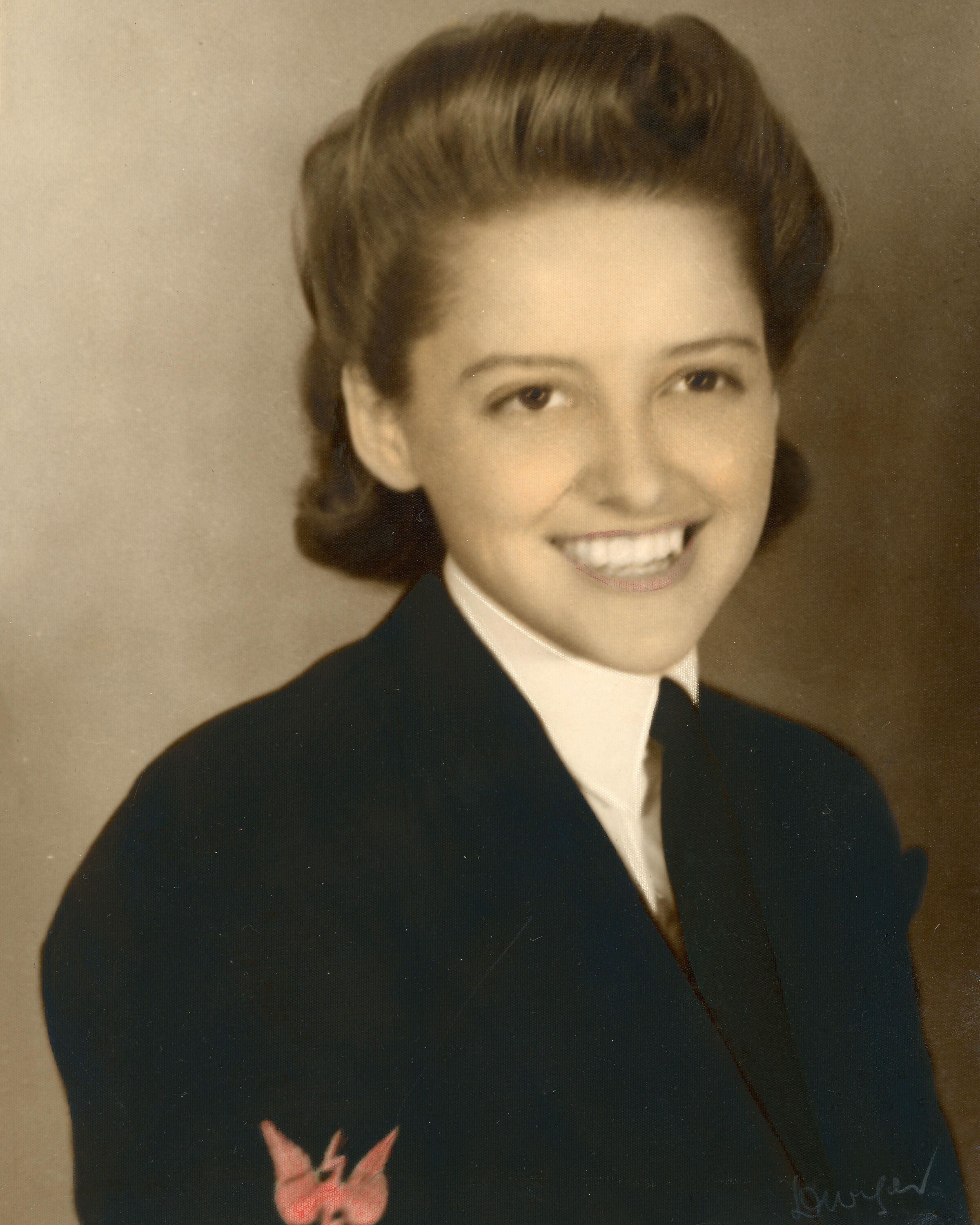 An old black and white photograph of a woman smiling, wearing a dark jacket and tie, and white shirt. There are hints of red where her lips and an insignia on the jacket sleeve have been coloured.  