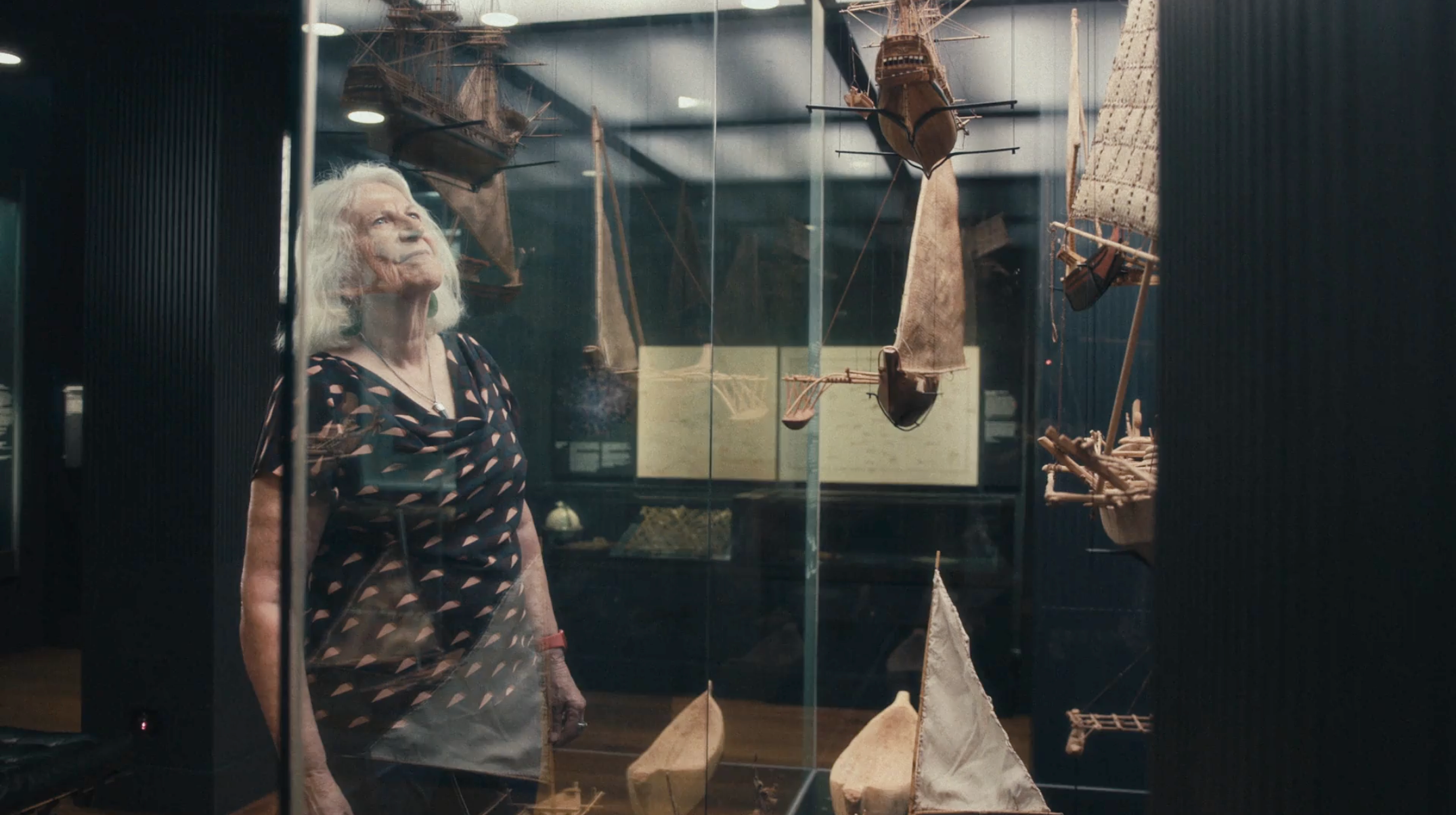Photo of a woman looking into a glass, museum display case featuring small boat models.