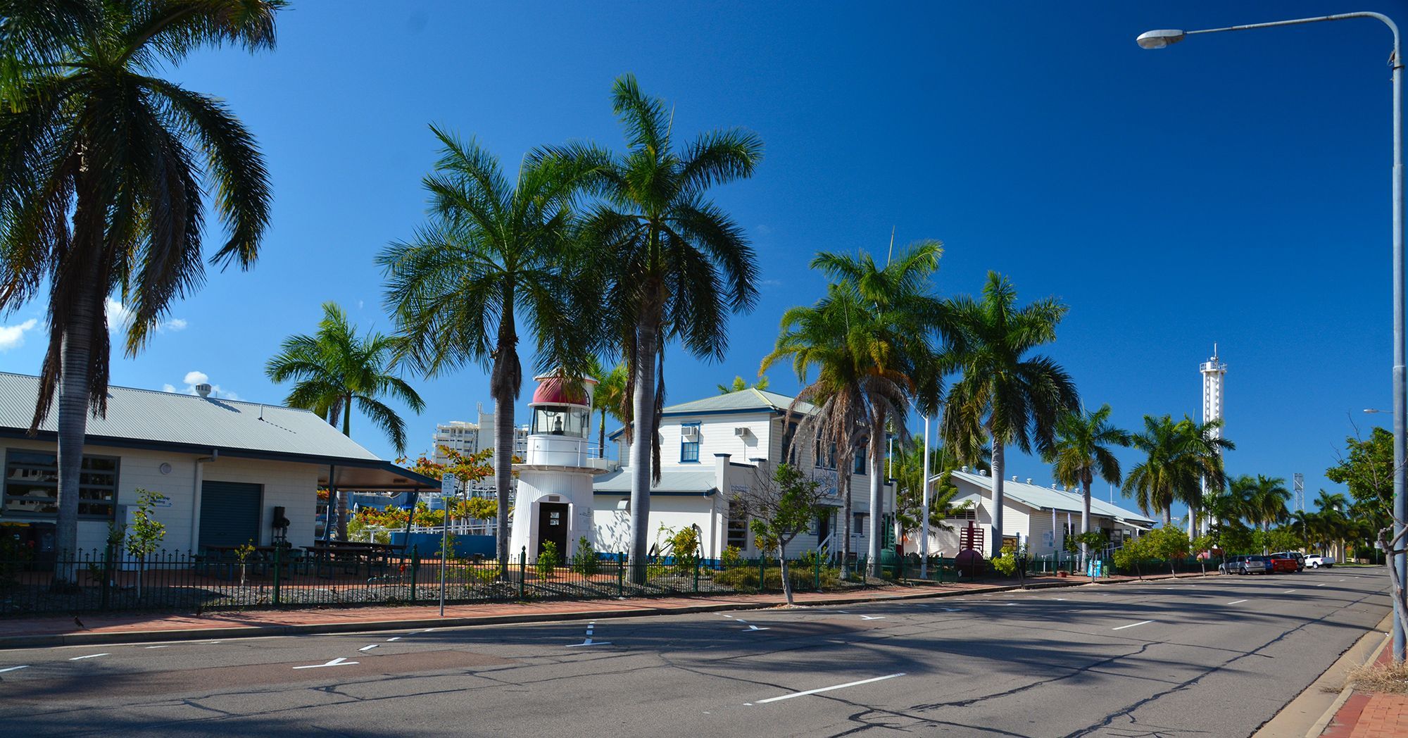 Photo showing a street lined with palm trees, with white buildings and a blue sky.