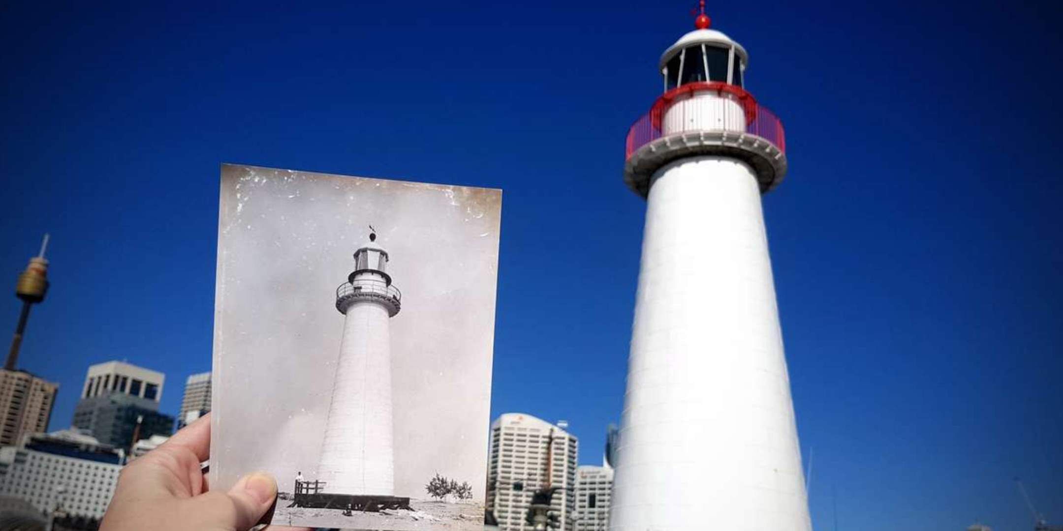Photograph of a hand holding up a black and white photograph of a lighthouse, infront of an actual lighthouse, white with red trim and a bright blue sky.