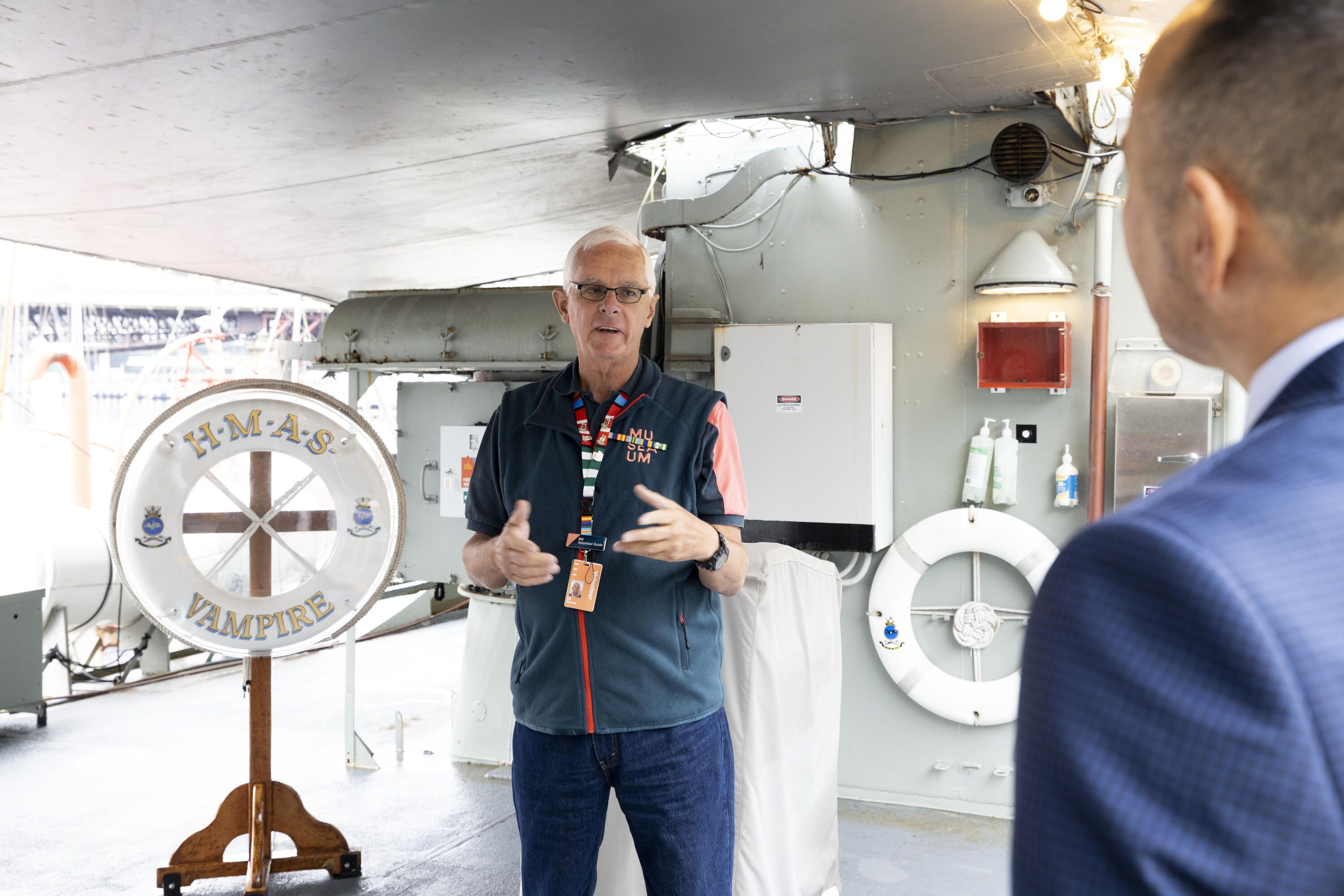 Photo of a male in a navy volunteers uniform standing on the deck of navy ship, the the shoulder of a man in the audience on the right side of the image.