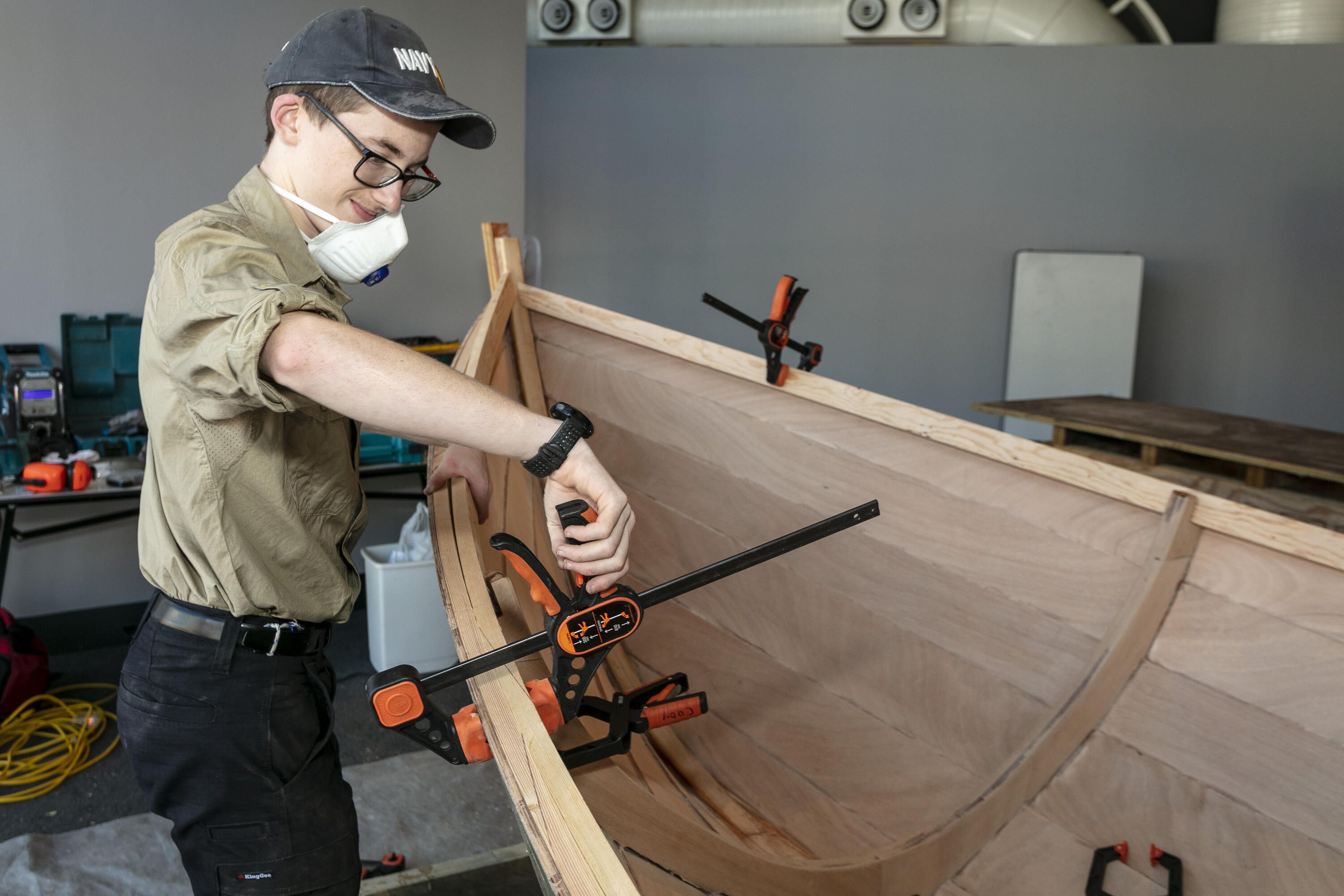 Photo of a teen boy fastening a clap on a wooden boat under construction