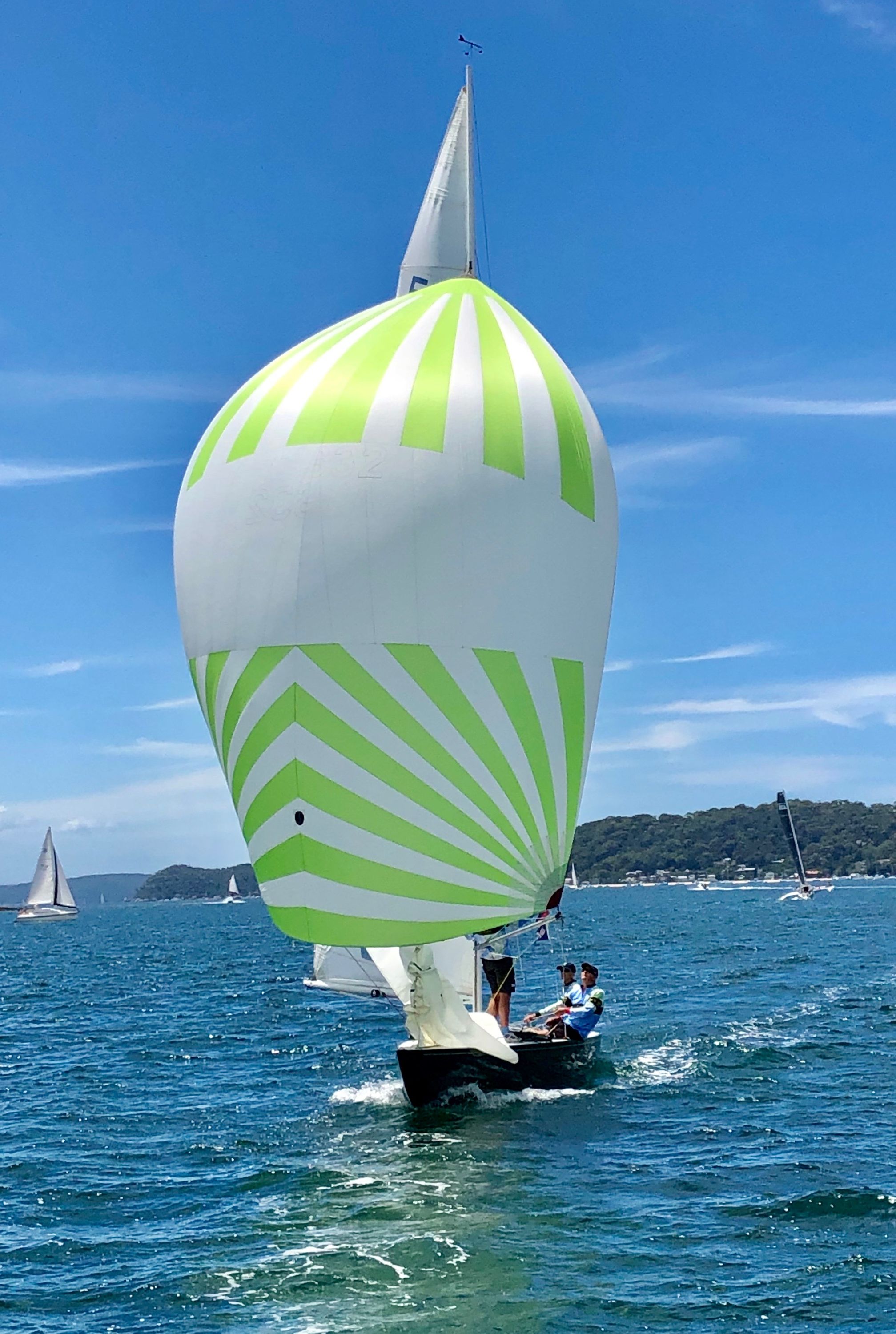 Photo of a boat sailing towars the camera, it's white sail with a green stripped pattern filled with wind.