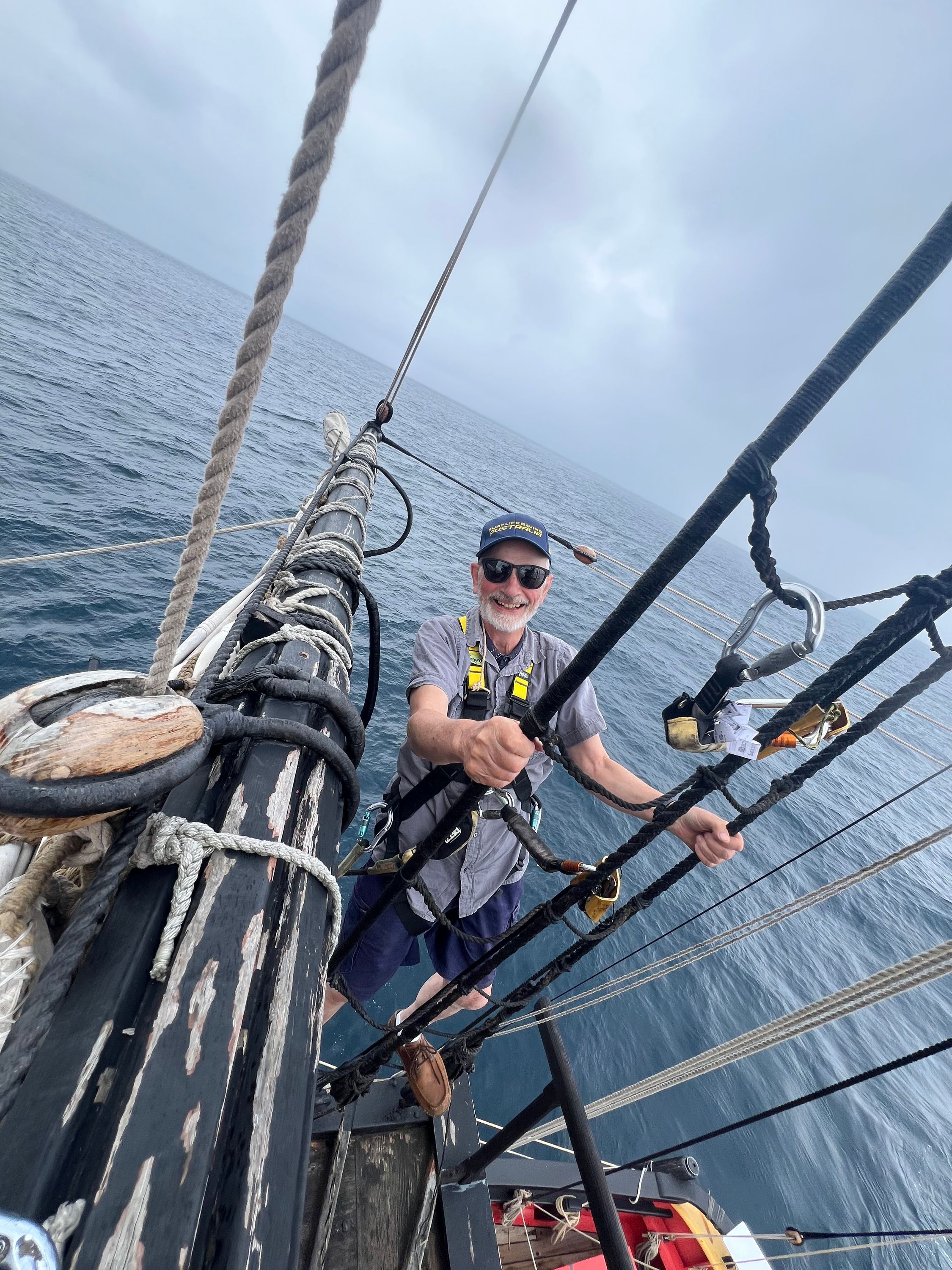 Photo of a man wearing a harness up the mast with the ocean behind him.