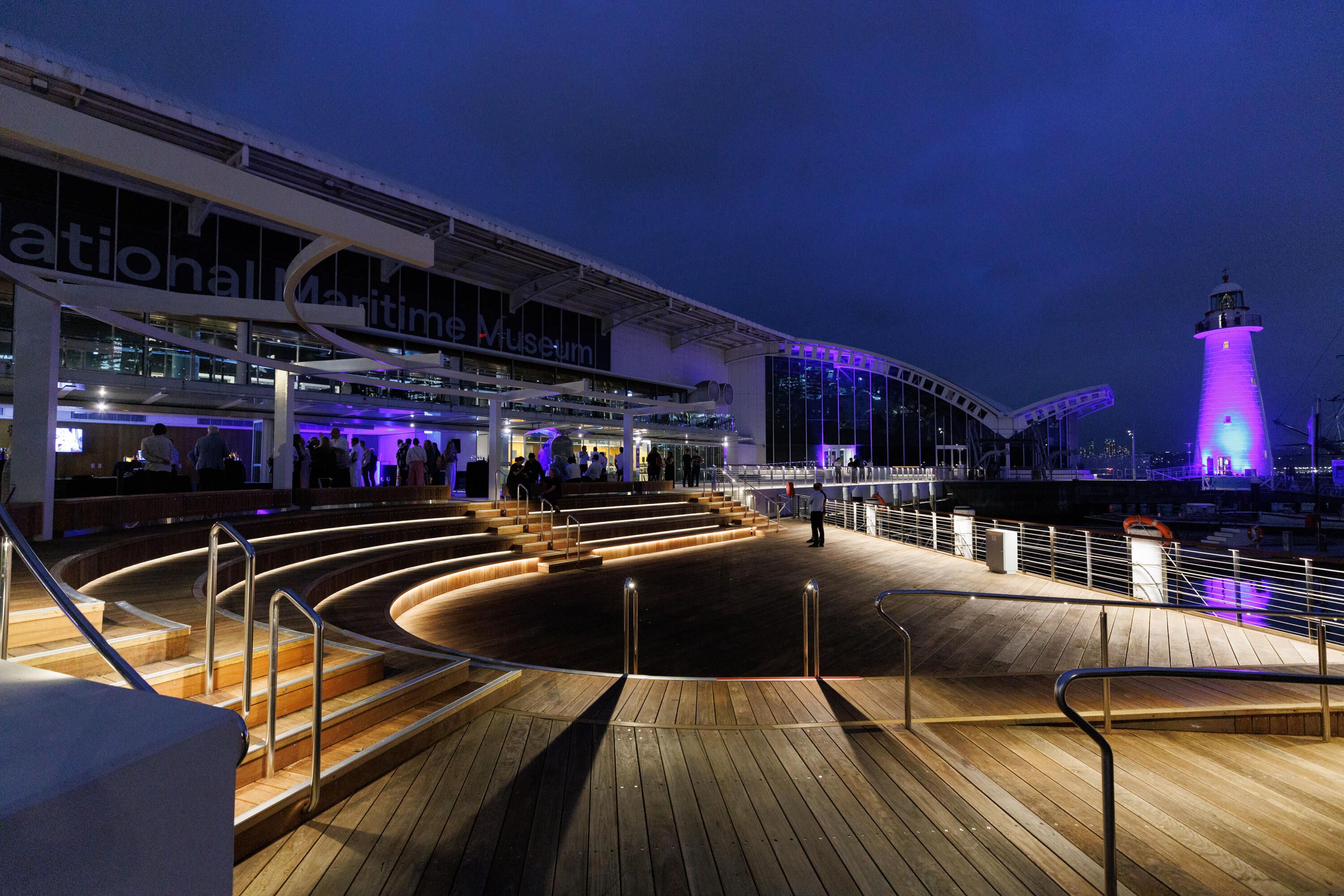 Museum waterfront at night. A seating area with lighthouse in the background. 