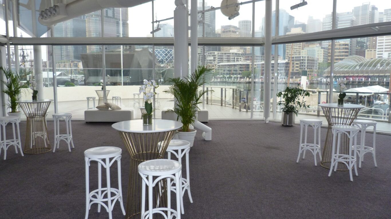 photo of an interior space with cocktail tables, stools and plants, with a full hight windows in the background.