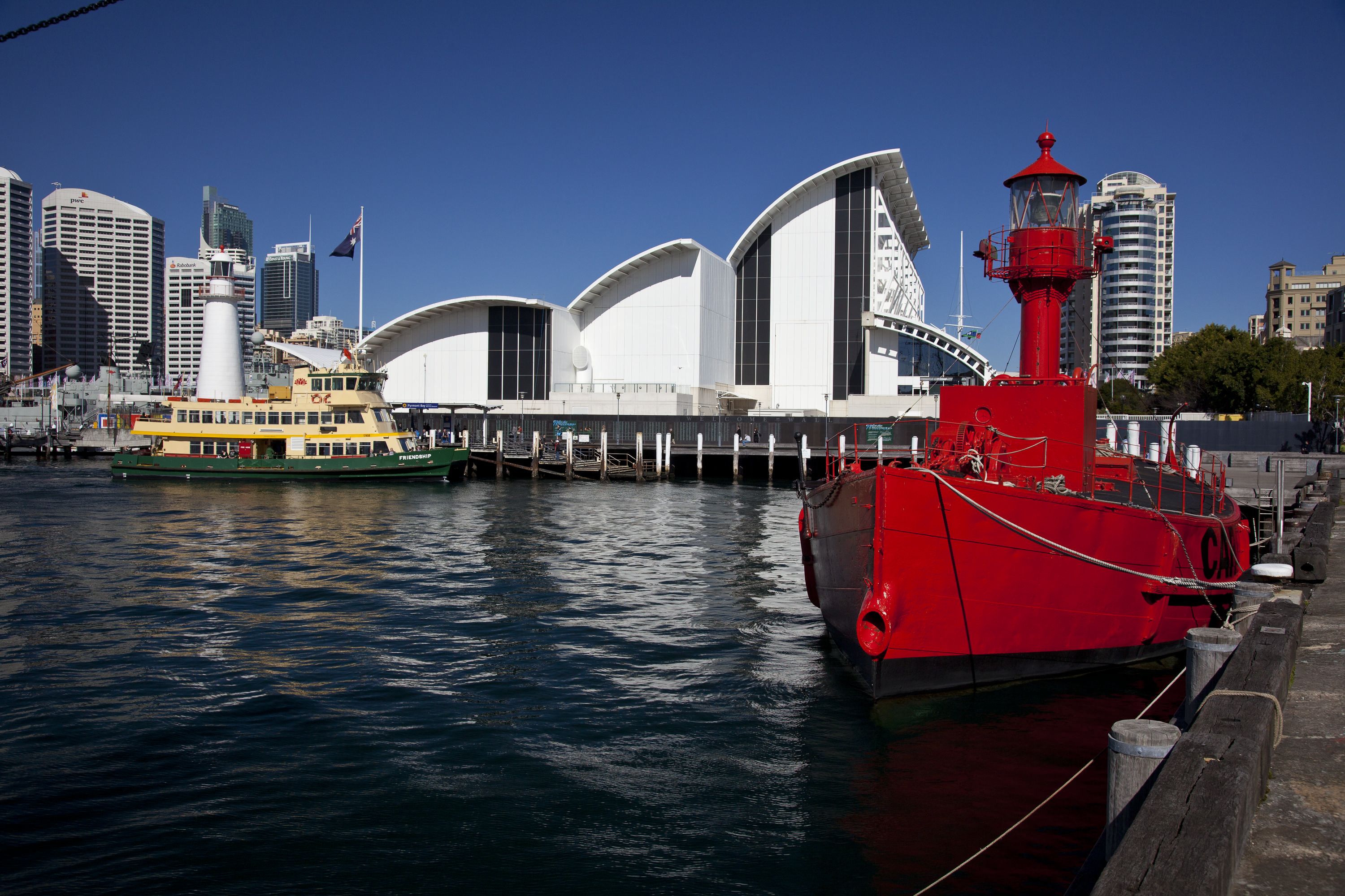 Photo of a red lighthouse ship floating in the harbour. the museum building and a sydney ferry are in the background. 