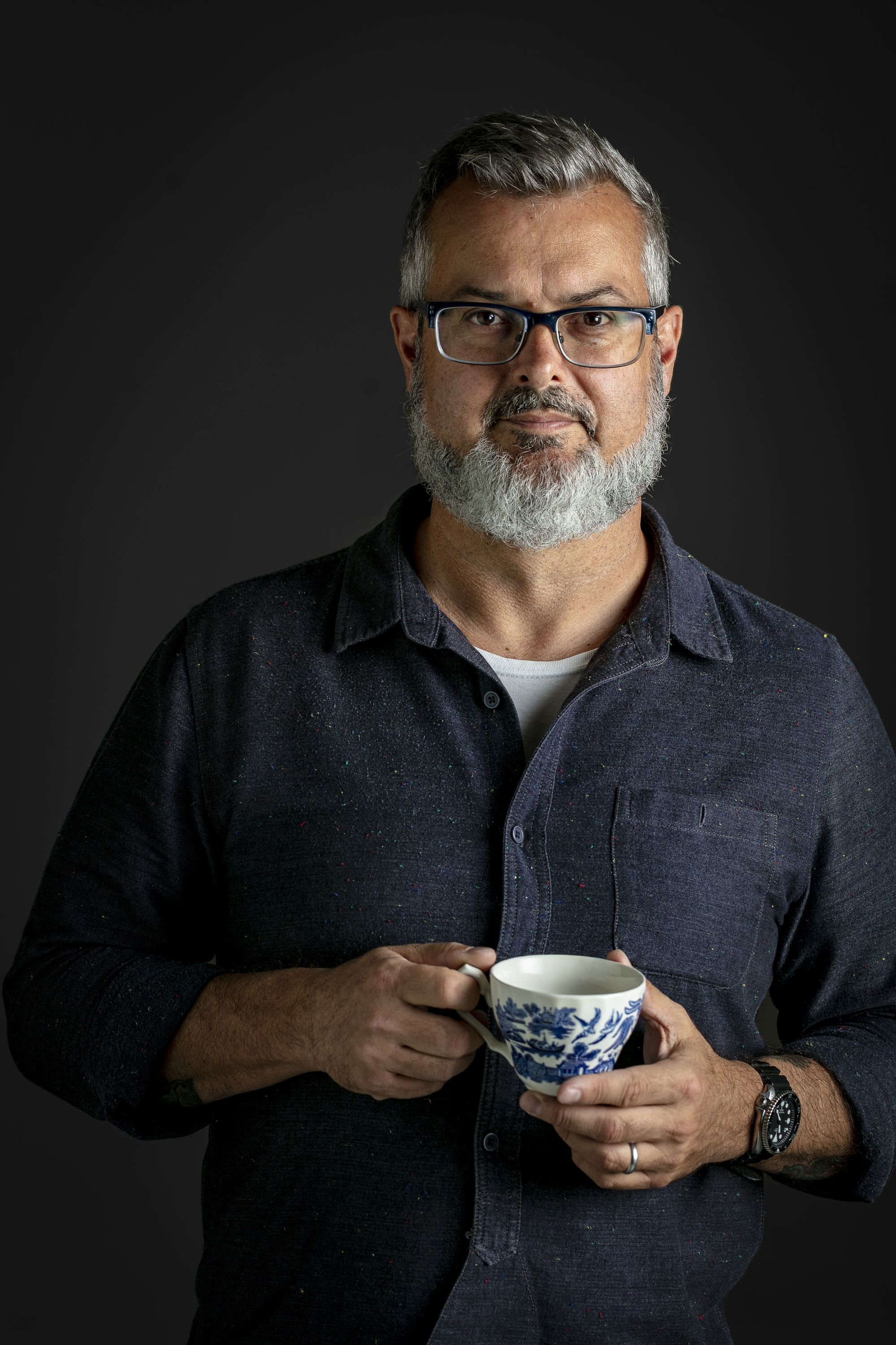Photo of a man with a grey beard, wearing a black button up shirt, holding a blue and white patterned teacup. 