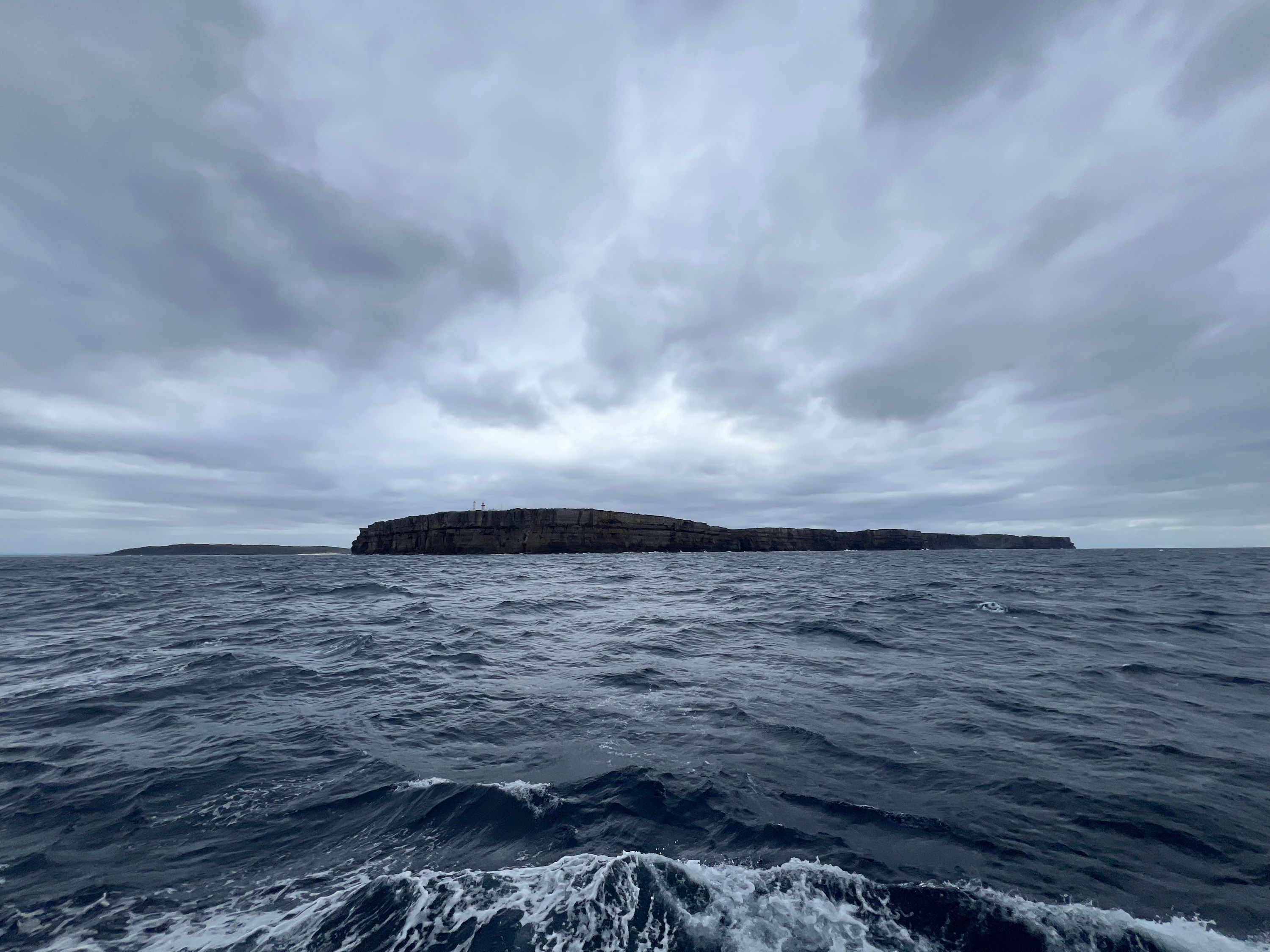 Photo showing a landscape, with a moody grey sky over a dark sea, with land in the middle of the horizon. 