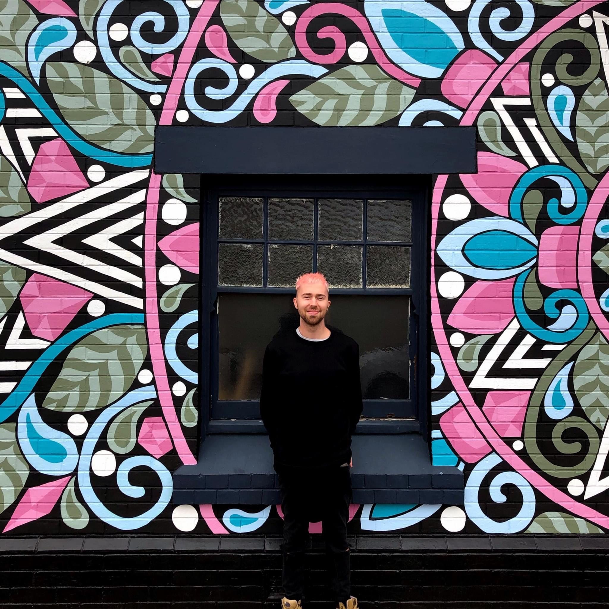 Photo of a man with blonde hair and clack clothes, standing infront of a building 