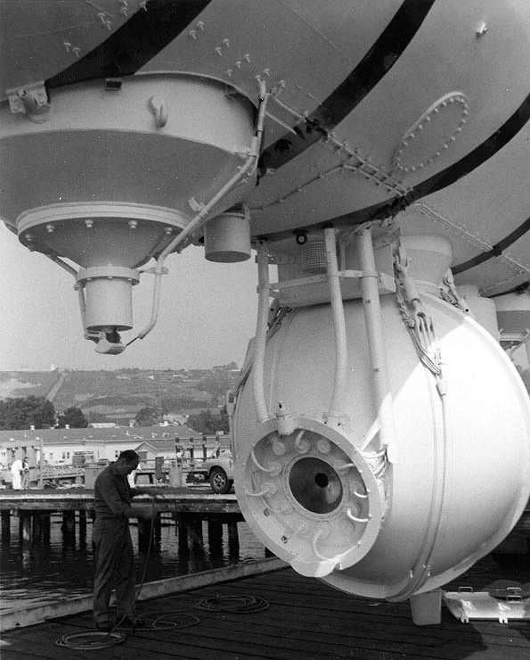 Black and white photo showing the underside of a submersible, with a large sphere only slightly larger than the man standing near it.