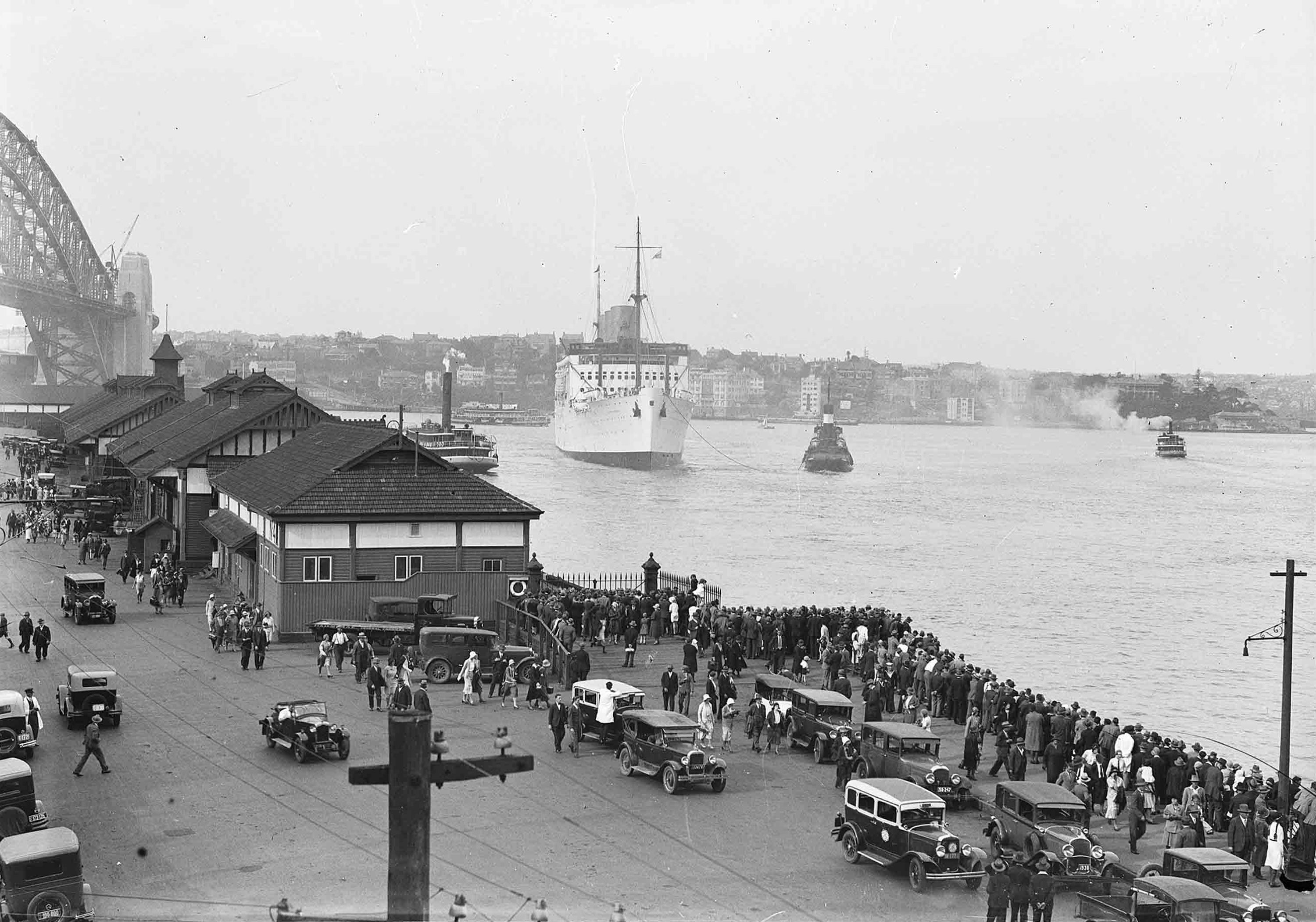A black and white photograph showing a large, white cruise ship sailing towards a dock where there are crowds of people and cars waiting for it. The Sydney Harbour Bridge is at the left edge of the image.