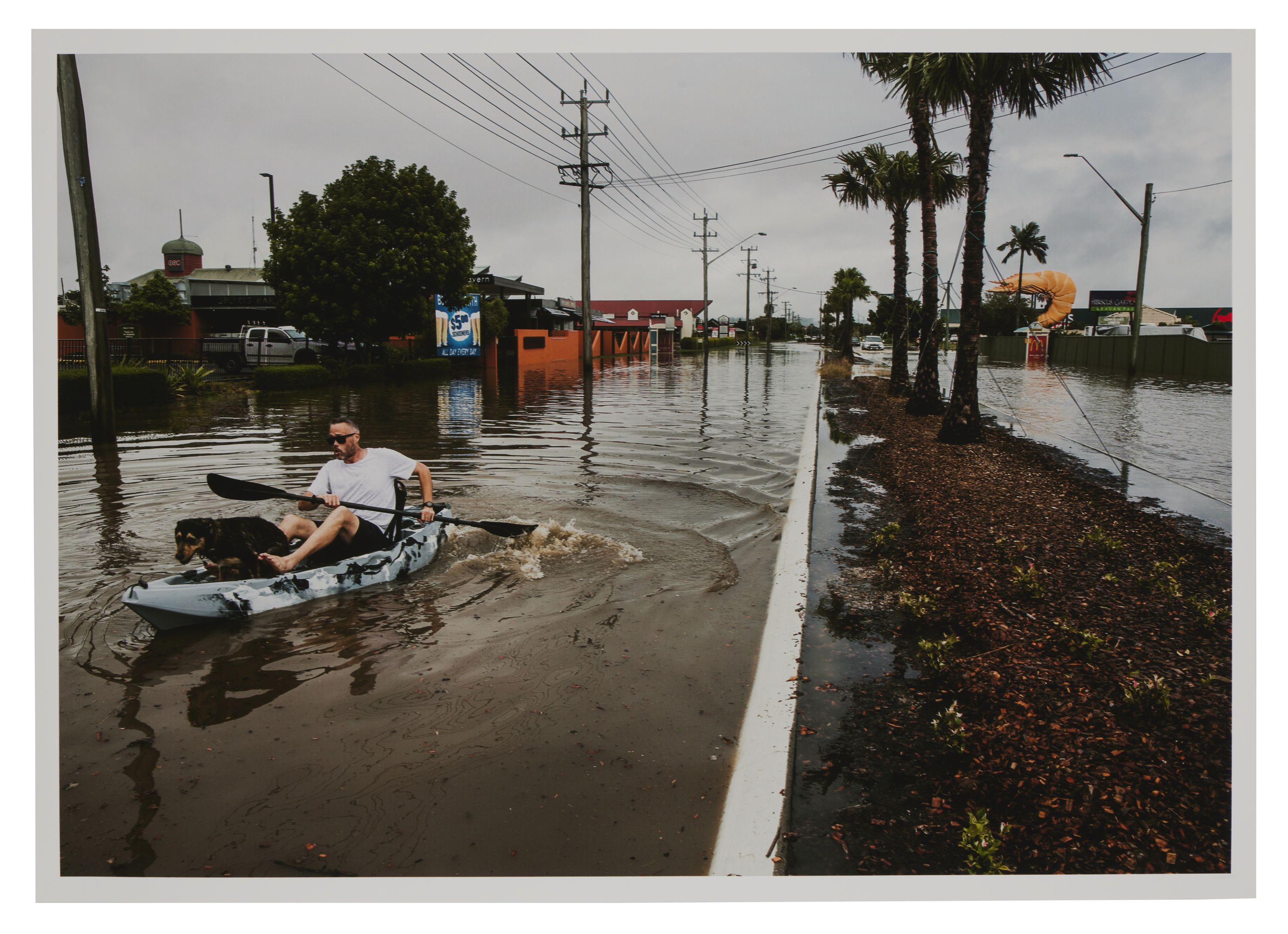 Photo of a street covered by water and a man on a paddleboard. 