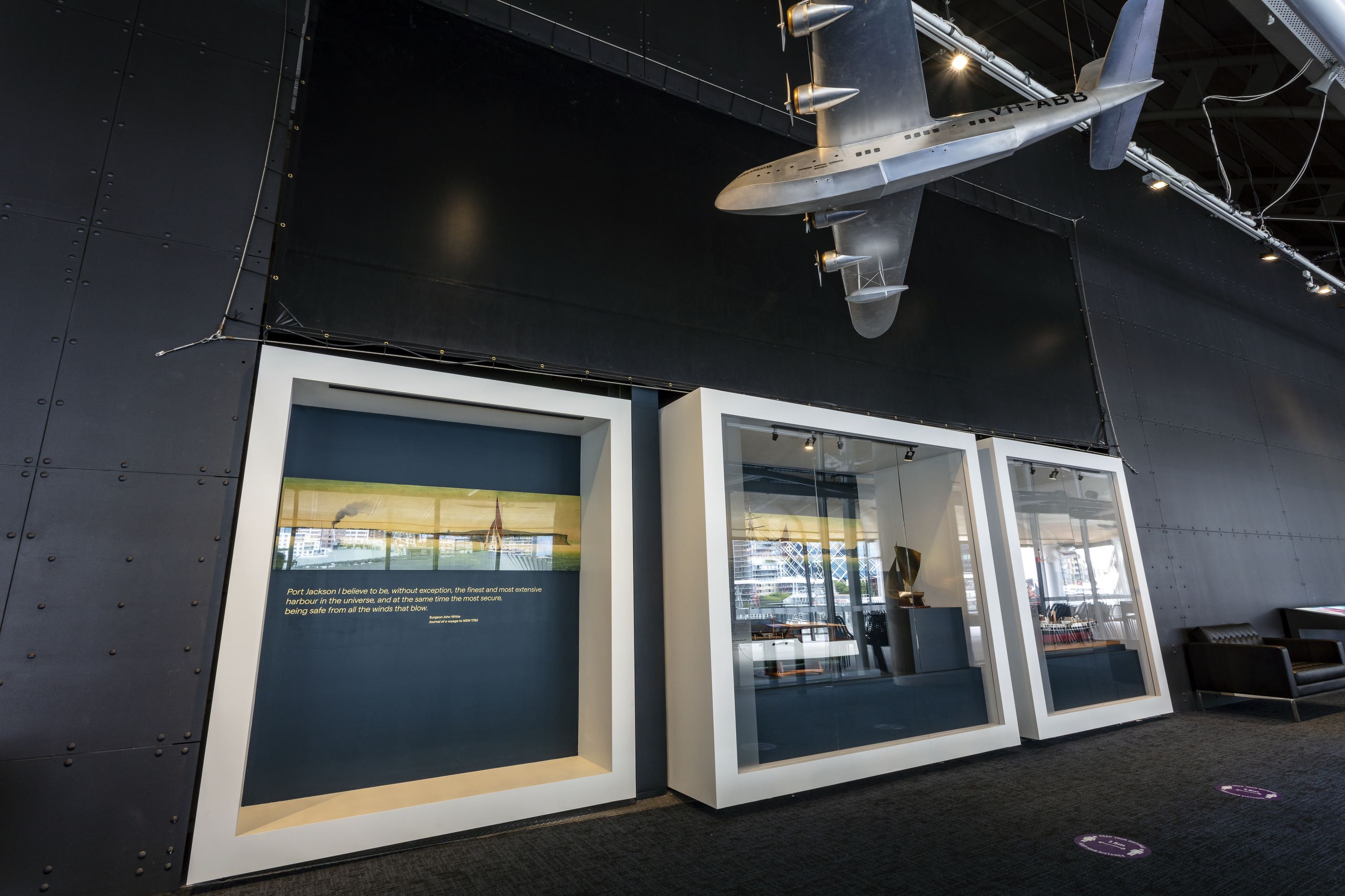 Photo of a museum exhibition, with display cases on the wall containing model boats and a silver model plane hanging from the roof. 