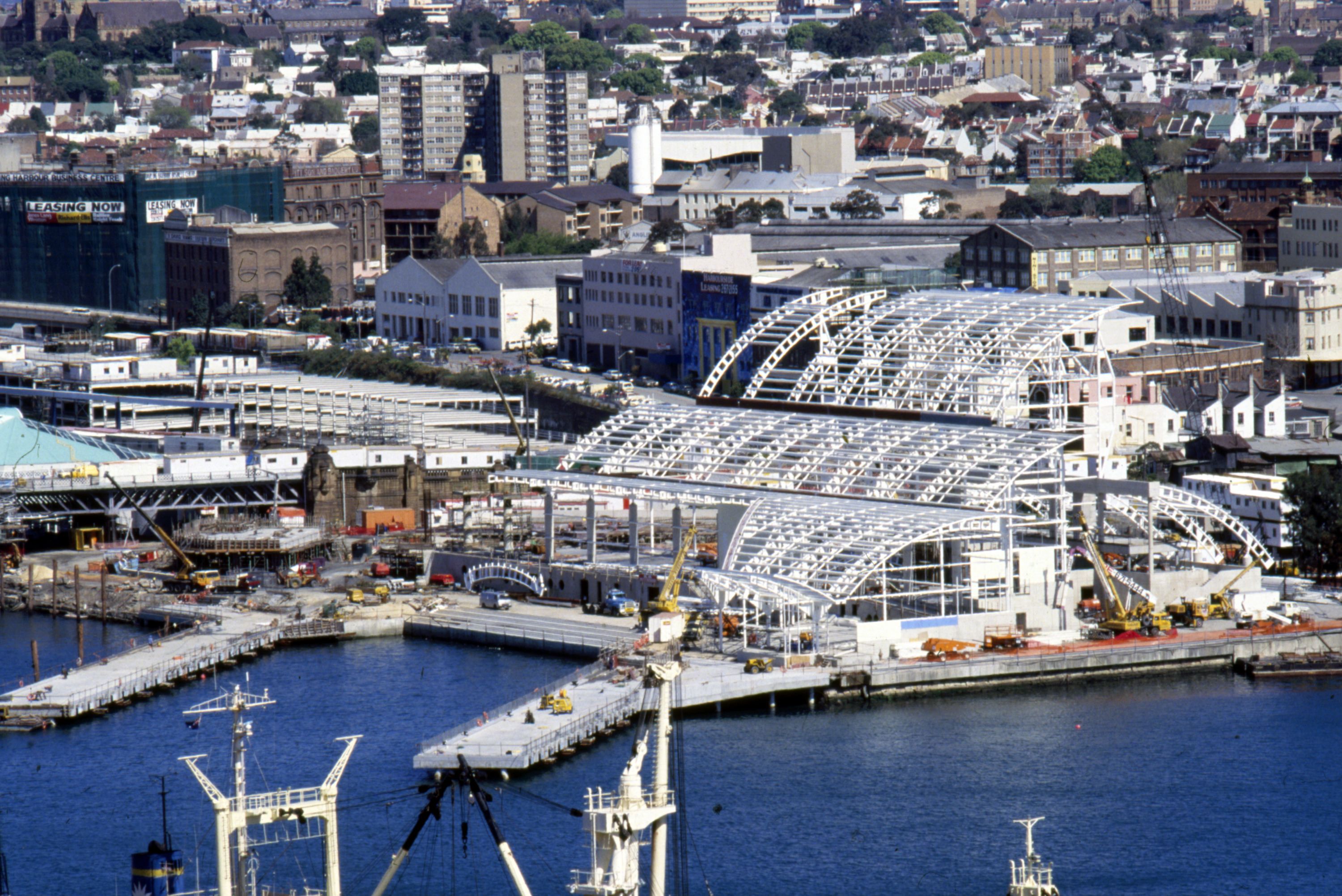 Aerial photograph showing a large building under construction, alongside the harbour. 